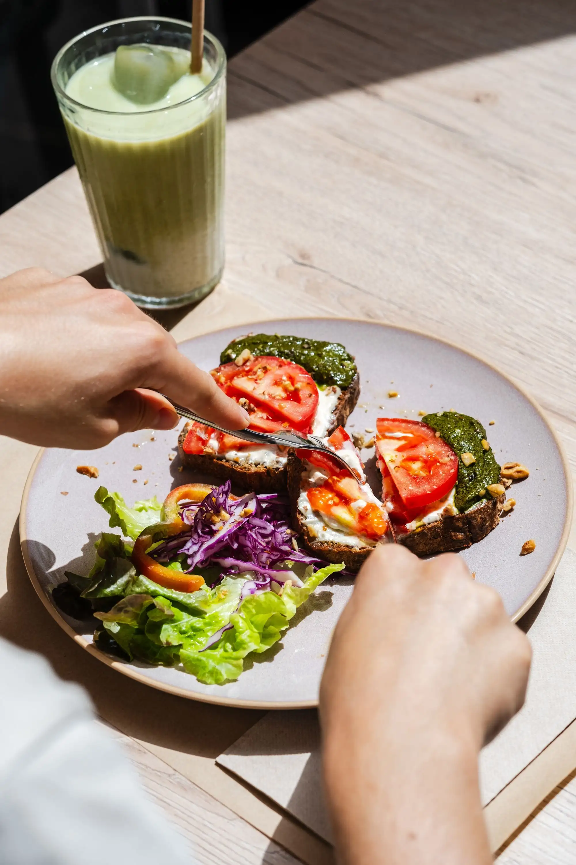 Hands cutting open-faced toasts with tomato and pesto beside a salad and a green smoothie