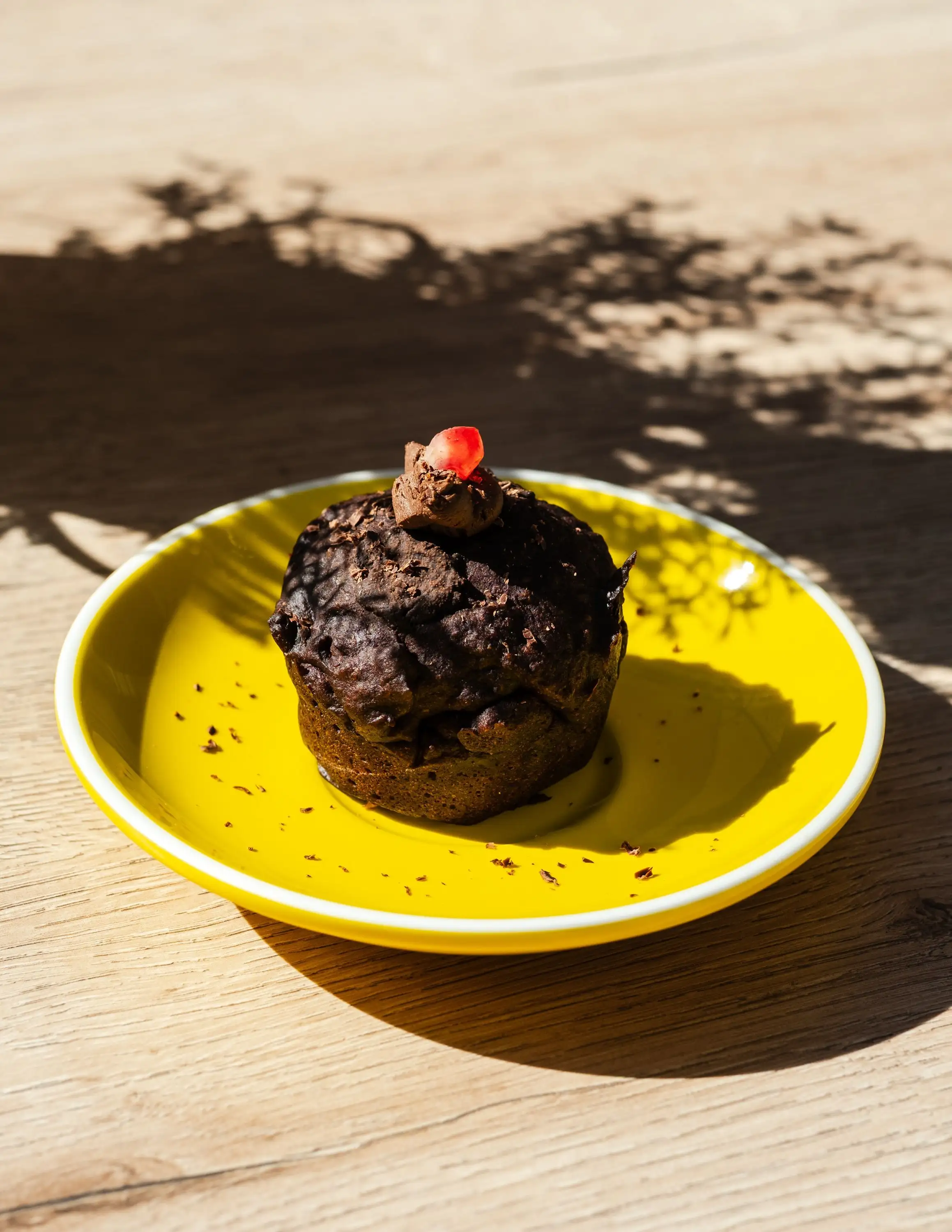 Chocolate muffin topped with a pomegranate seed on a yellow plate with leaf shadows on a wooden table