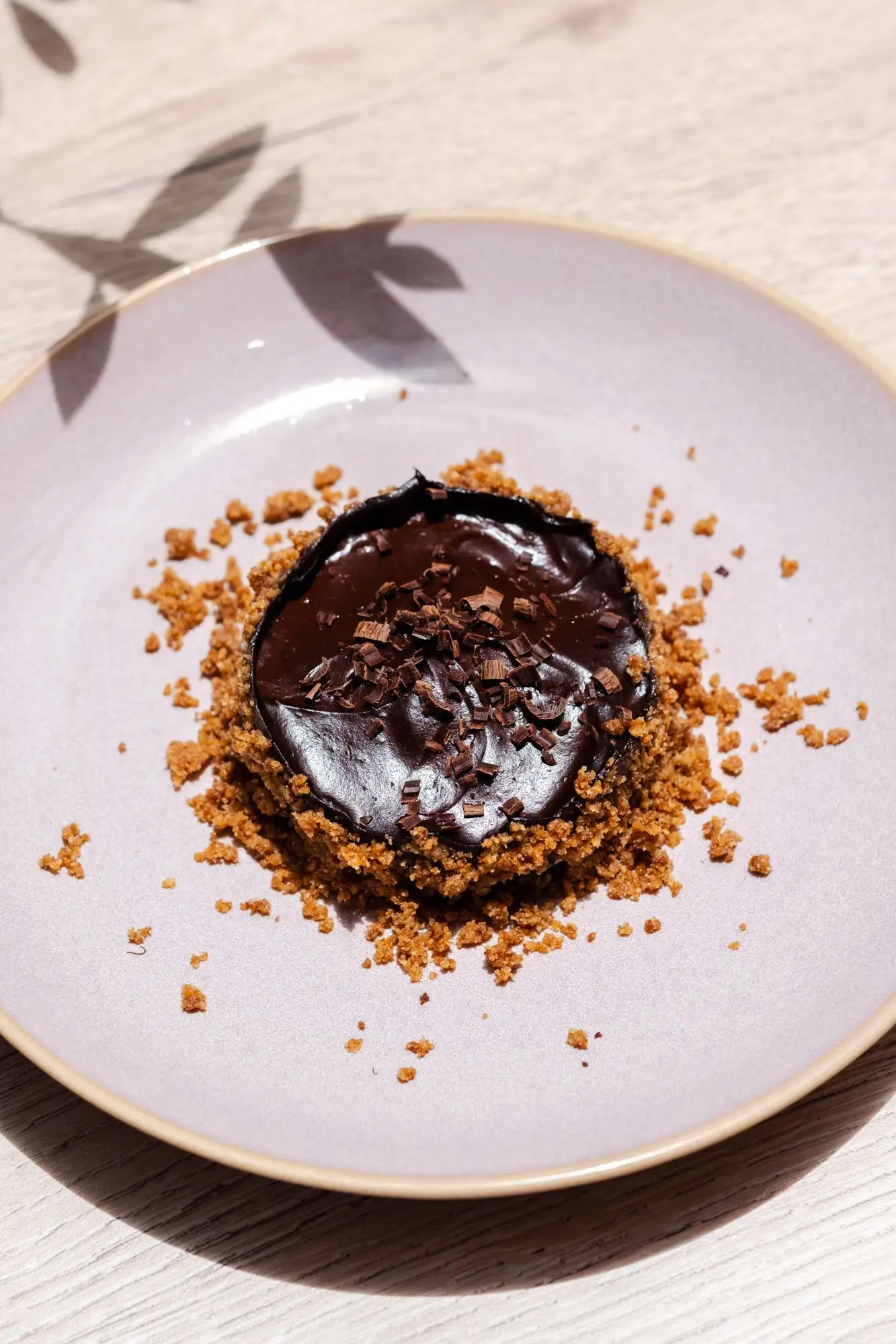 Chocolate tart with shavings on a pale plate surrounded by biscuit crumbs