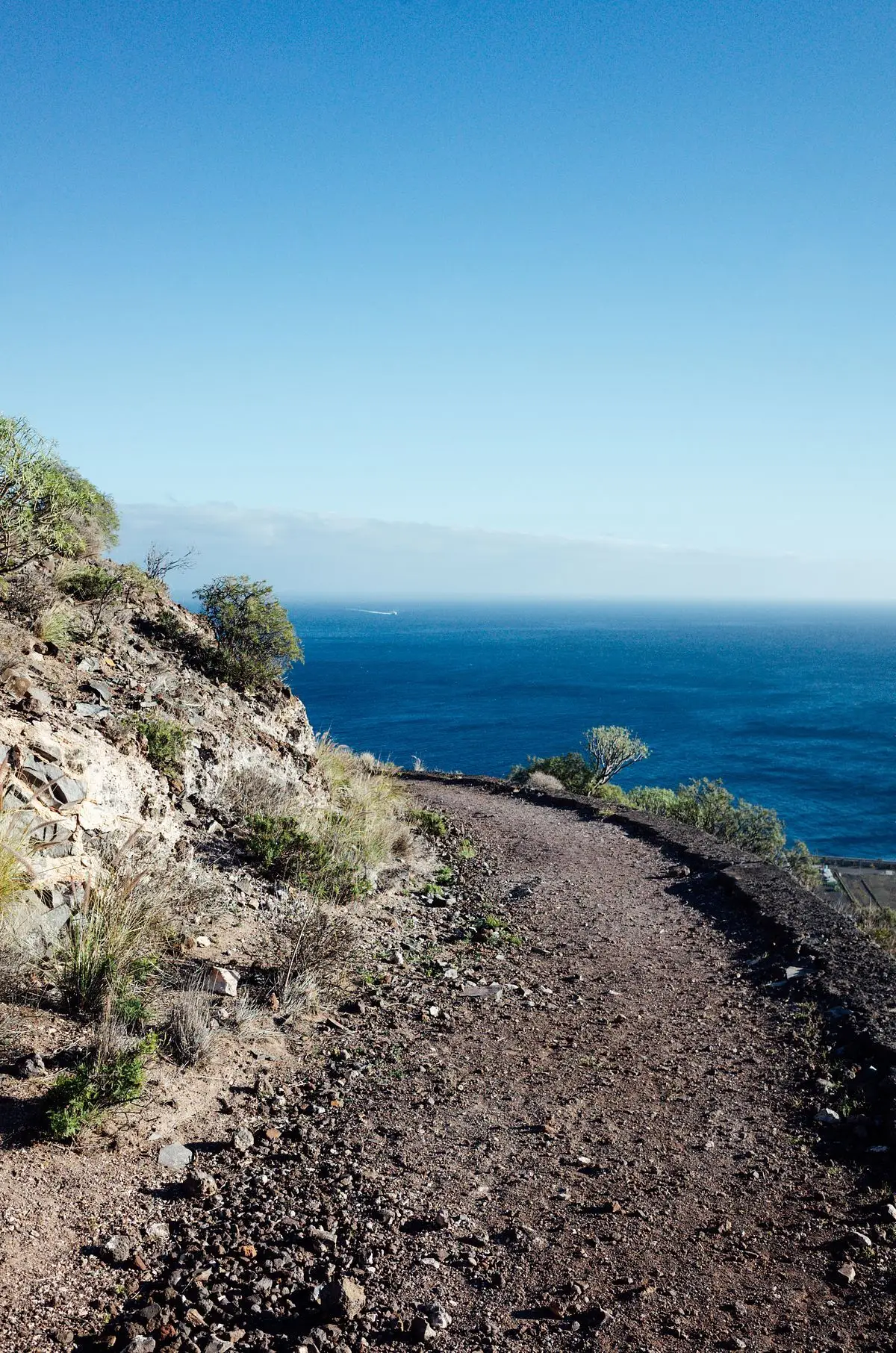 Sendero de tierra por una ladera seca sobre un océano azul intenso, con el cielo despejado