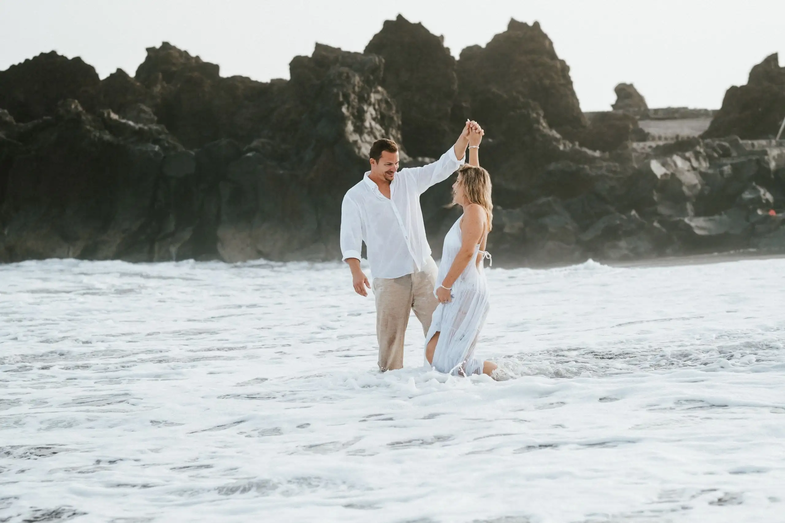 Man twirling a woman in white while standing in foamy surf in front of jagged black volcanic rocks