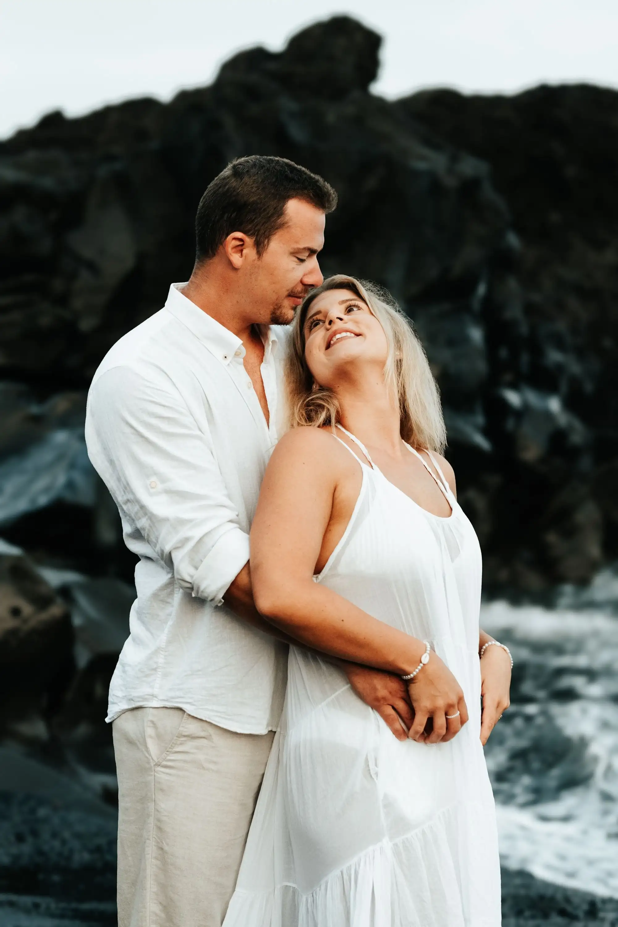 Man in white shirt embracing a smiling woman in a white dress from behind, dark volcanic rock behind them