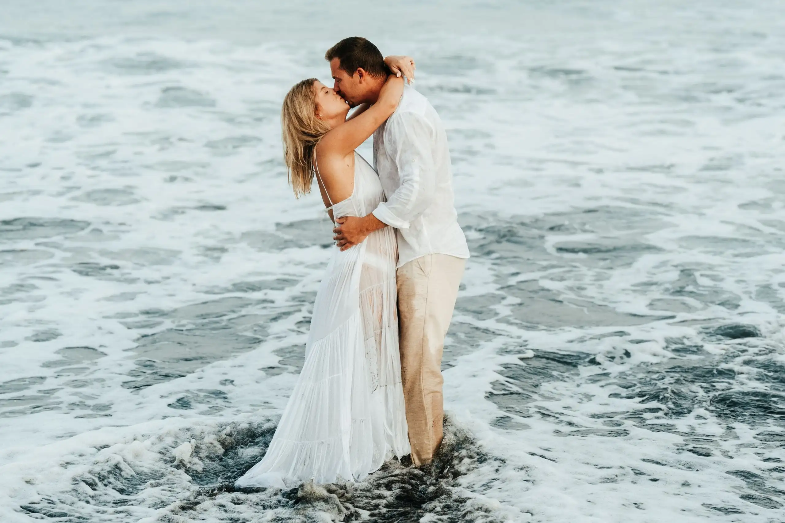 Couple embracing and kissing while standing in foamy surf around a dark rock