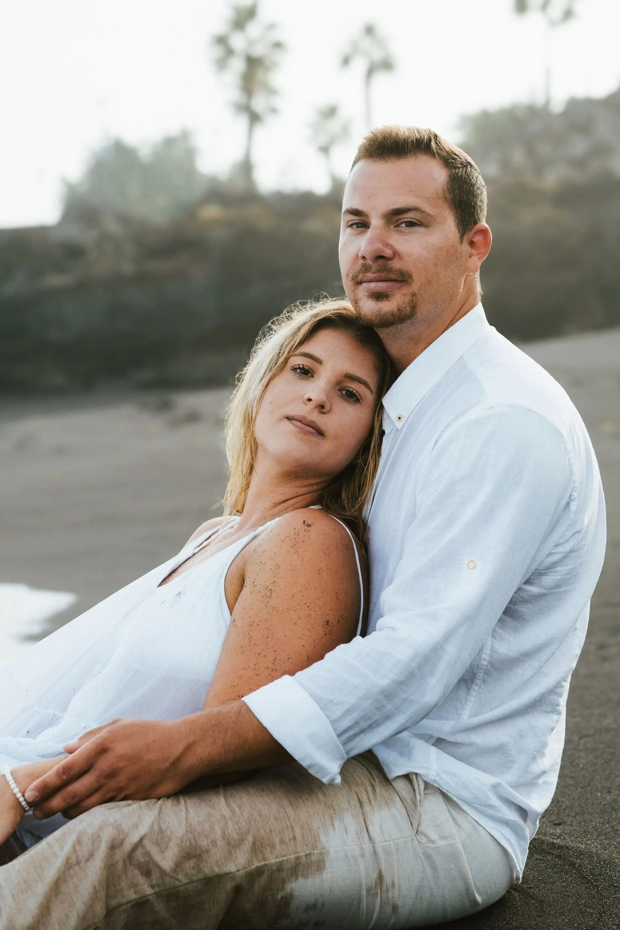 Woman leaning back against a man in a white shirt as they sit on black sand, palm trees blurred behind