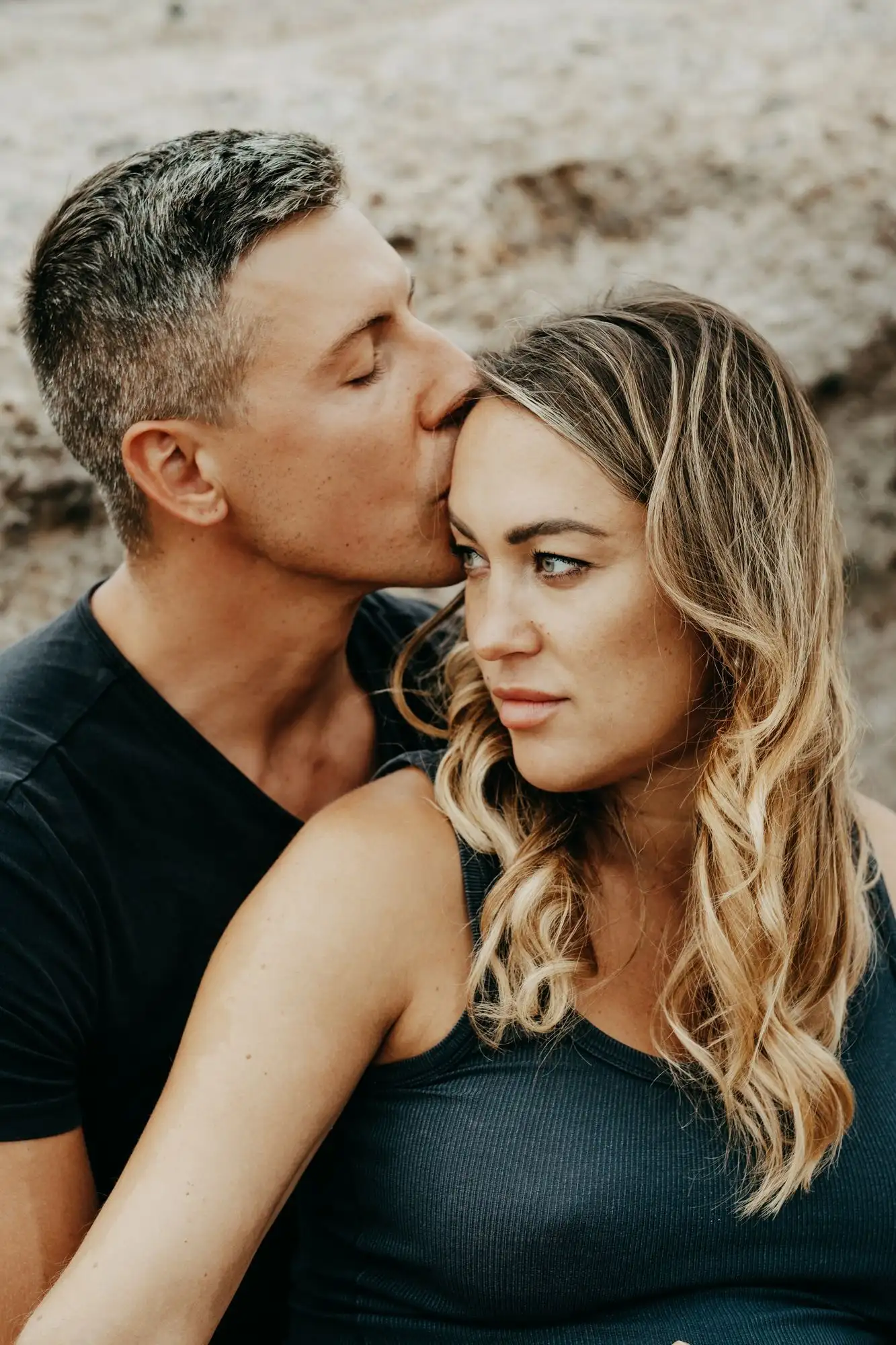 Close-up of a man kissing a woman on the forehead, both seated against a sandy rock wall