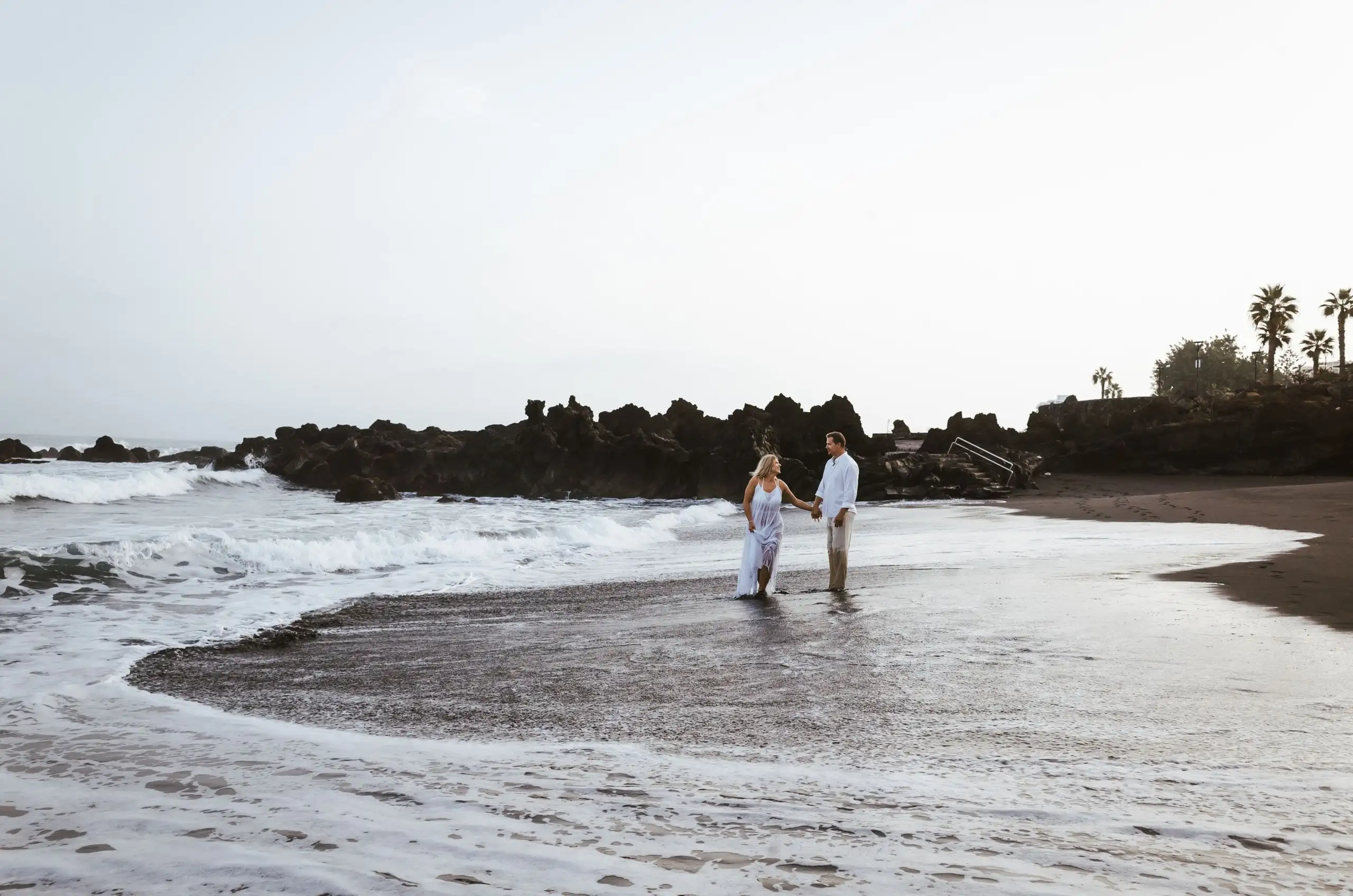 Couple in white clothes holding hands in the shallows of a dark-sand cove with volcanic rocks and palms behind