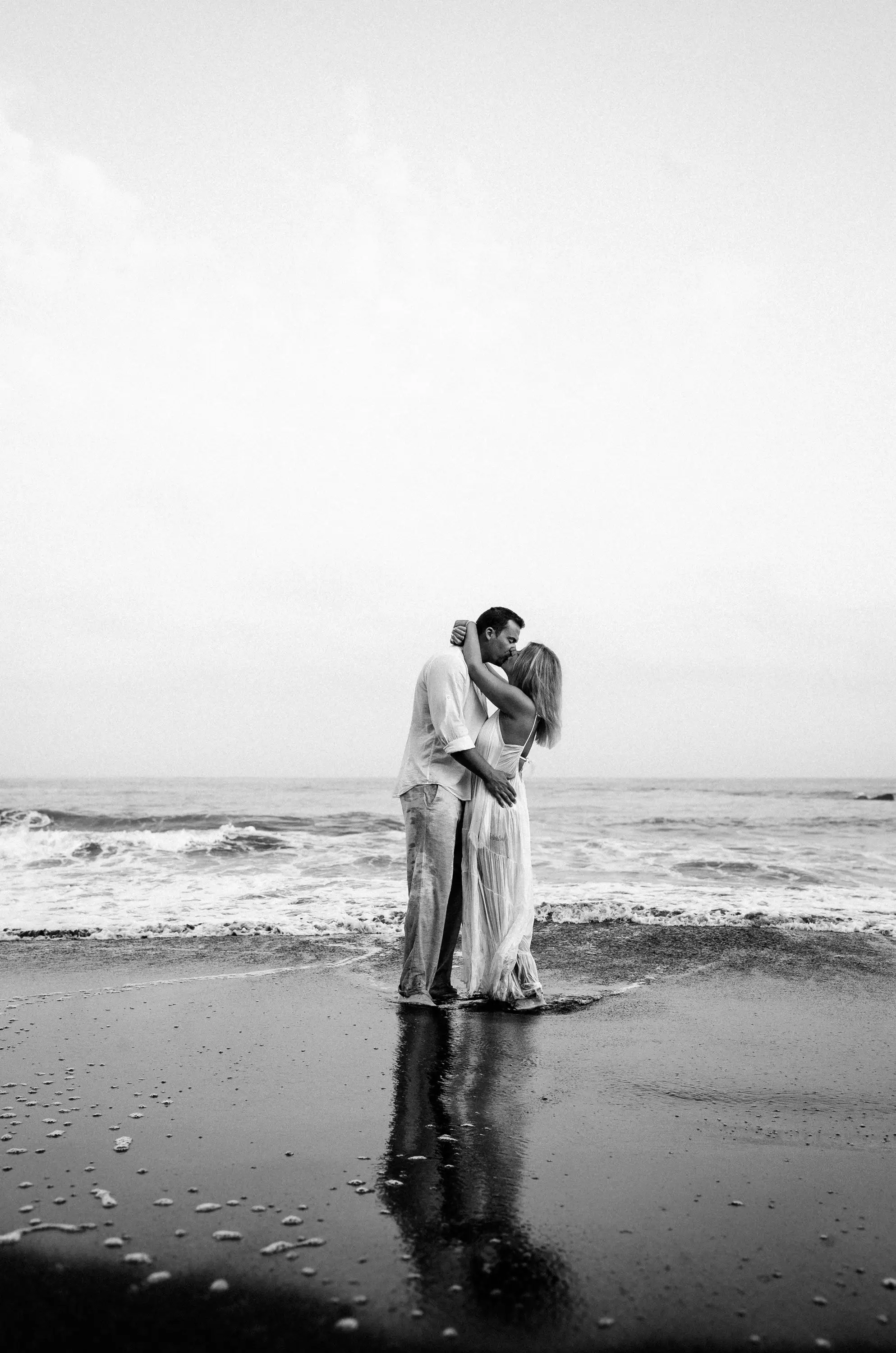 Black and white image of a couple kissing on wet black sand at the shoreline, reflected in the water