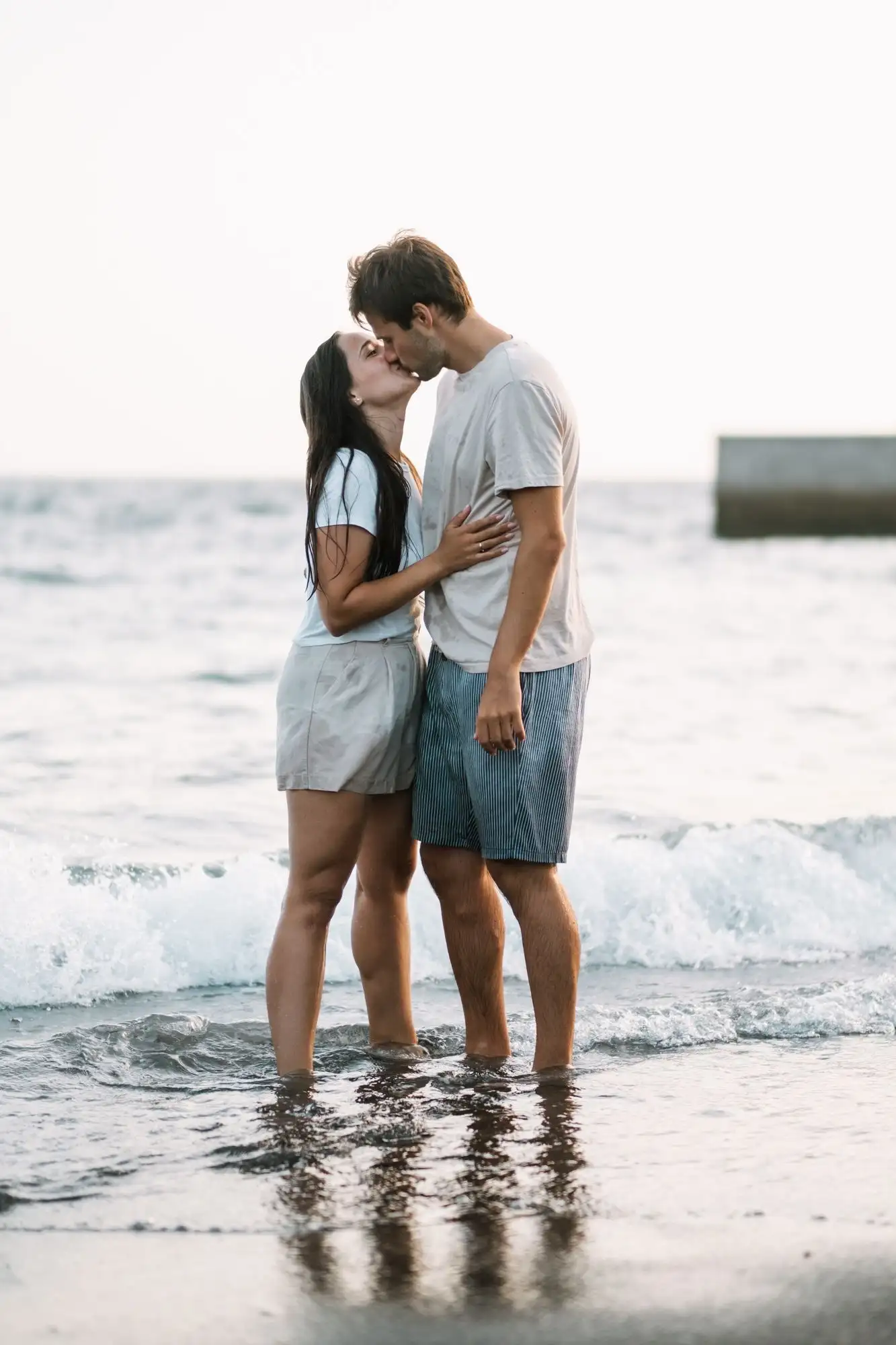 Couple kissing while standing ankle-deep in the sea as a wave breaks behind them
