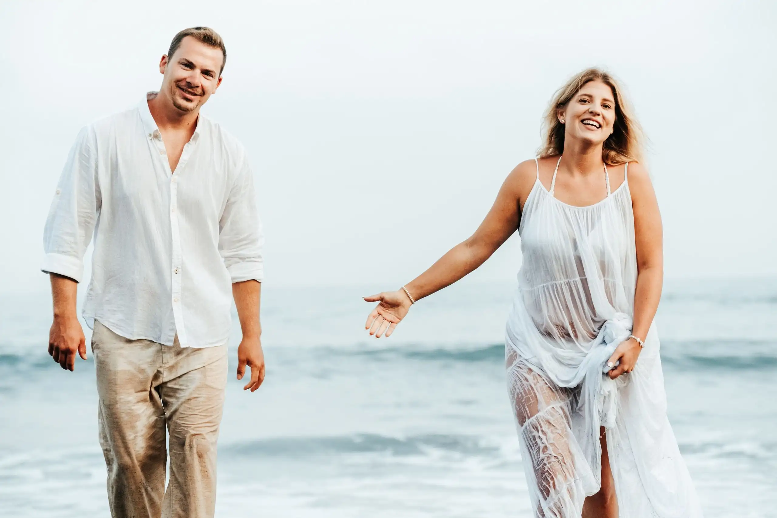 Laughing couple in white linen walking through shallow sea, the woman reaching out a hand