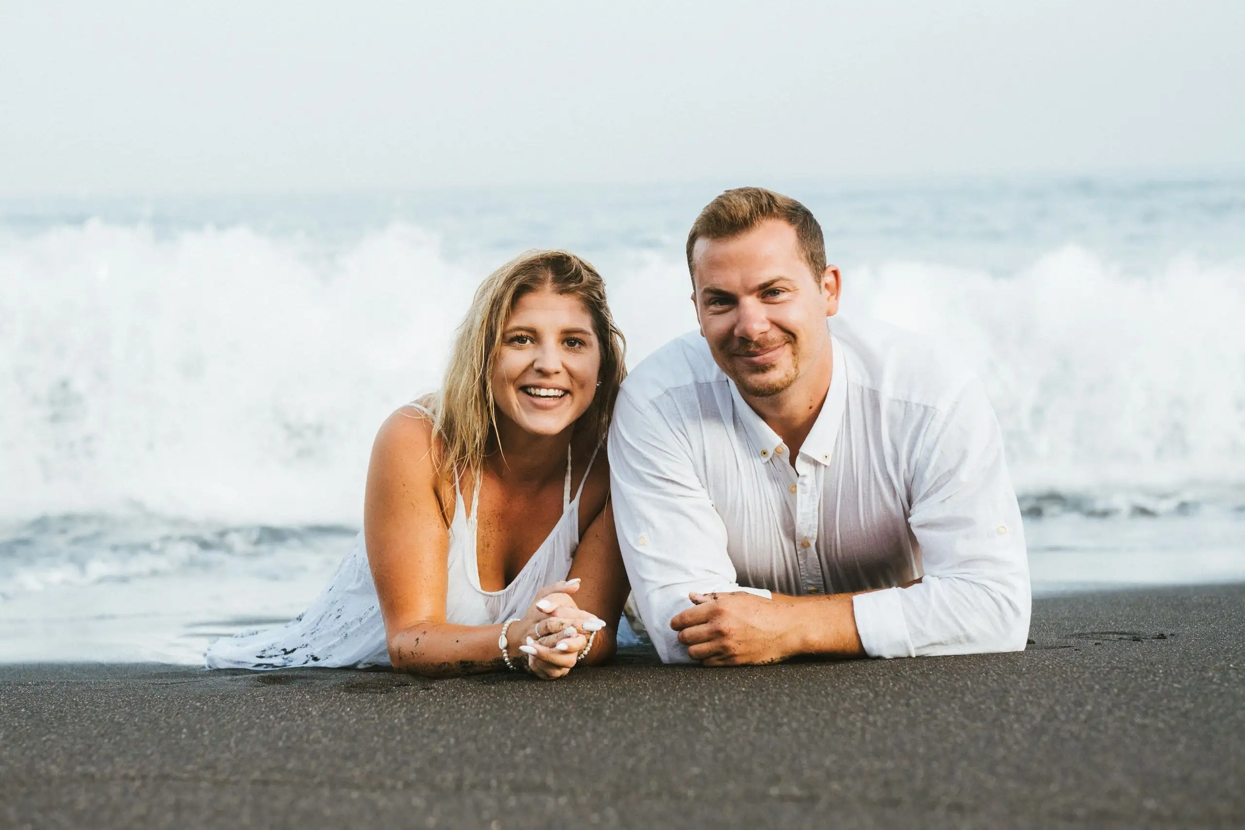 Smiling couple lying on black sand holding hands with a wave breaking behind them