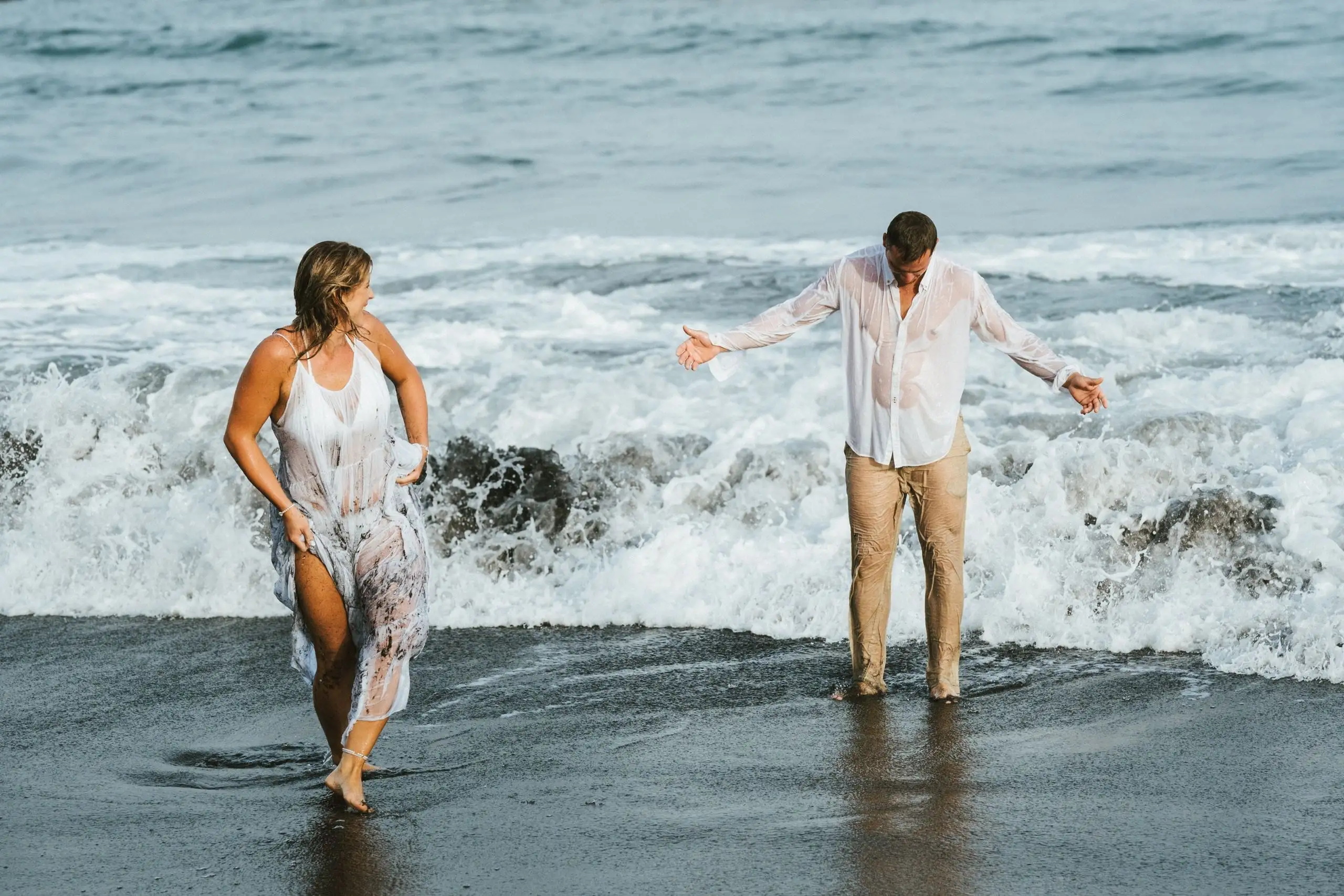Couple in white clothes playing in breaking waves on a dark beach, the woman walking out of the water