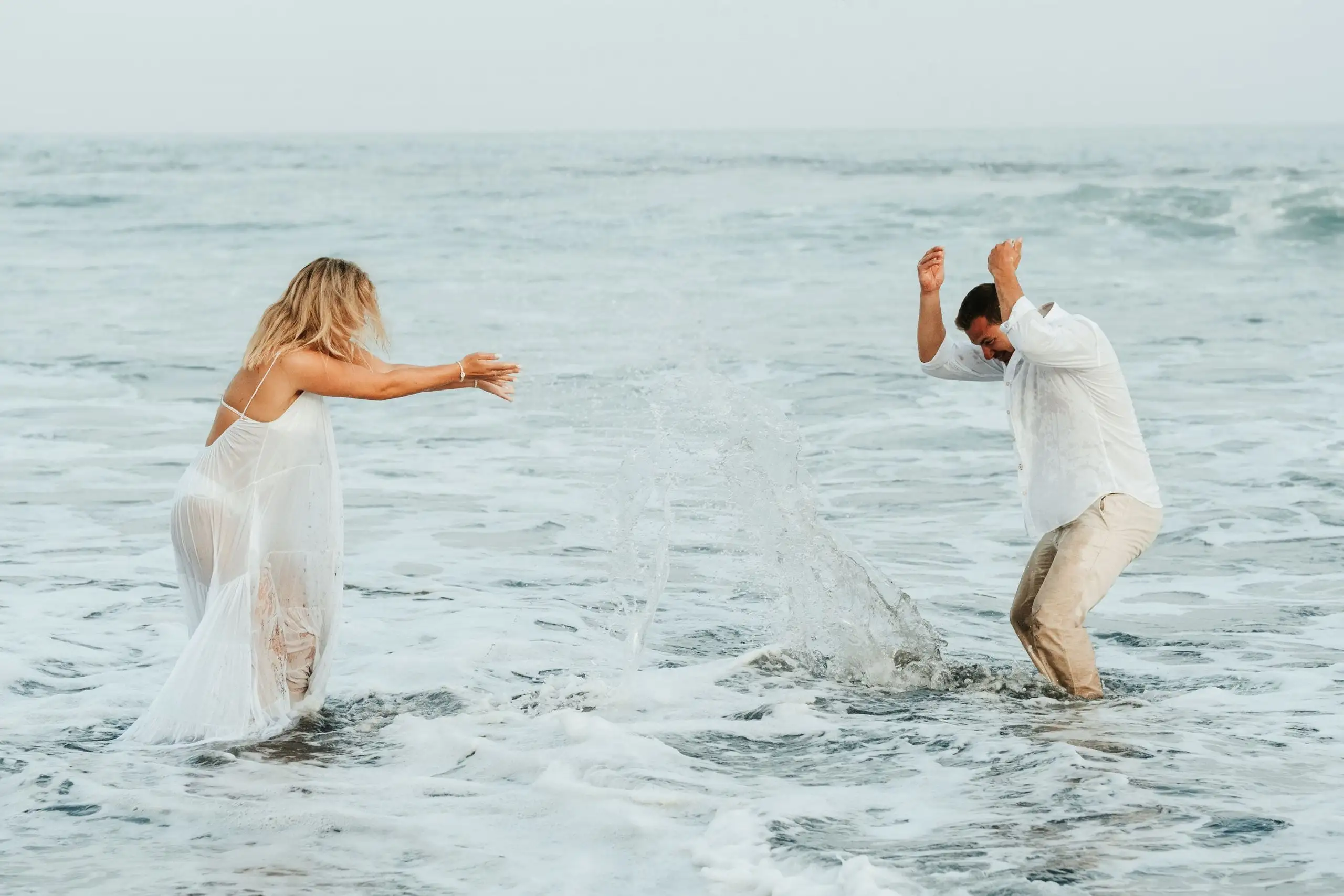 Couple splashing water at each other in the shallow sea, the woman in a white dress and the man in a white shirt