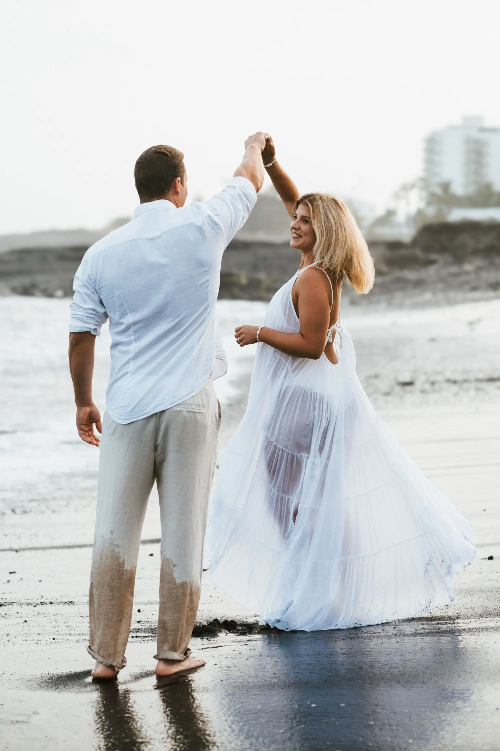 Man twirling a woman in a flowing white dress on wet sand by the sea, seen from behind