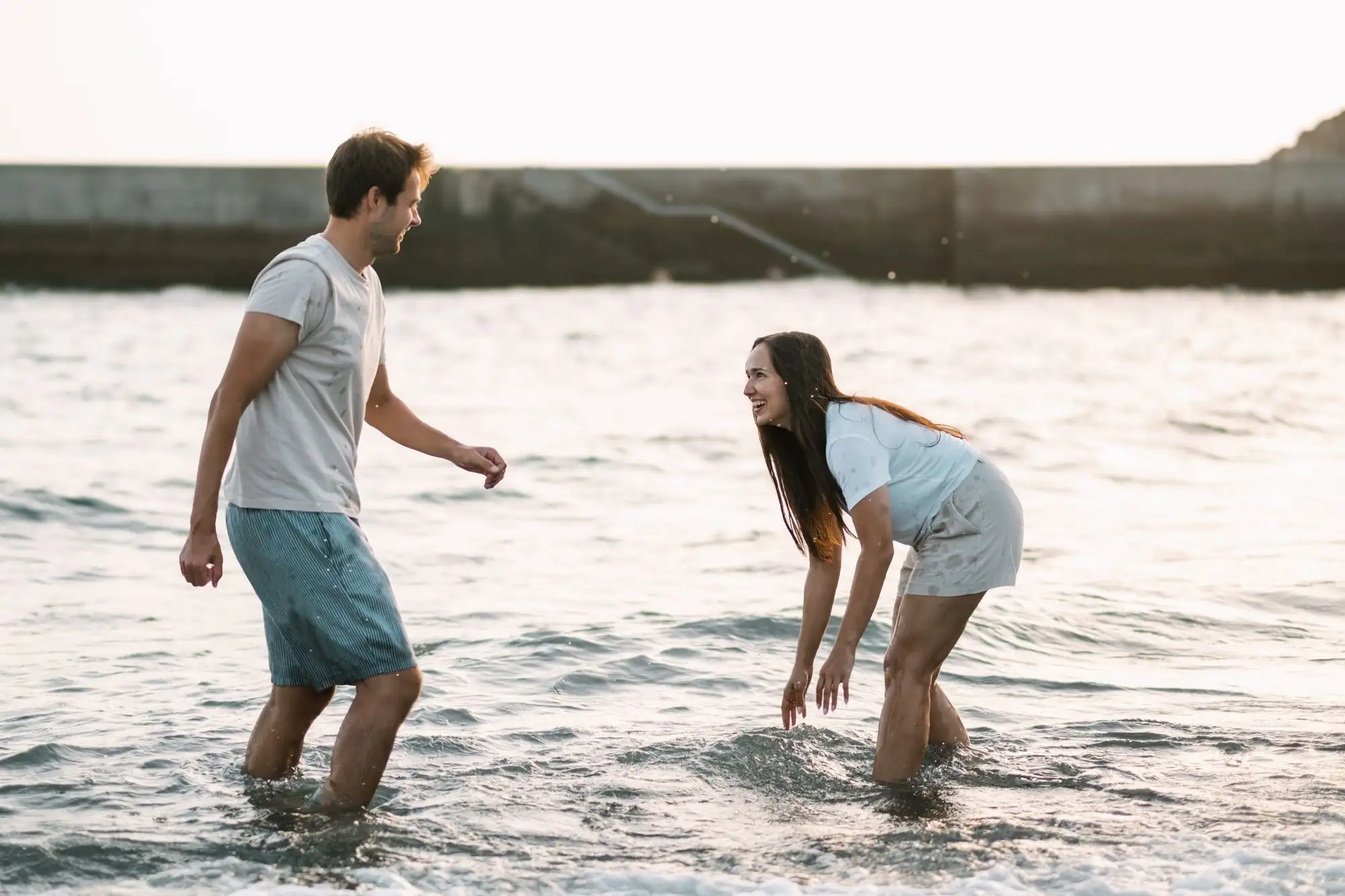 Man and woman laughing as they wade through shallow sea water in front of a harbour wall