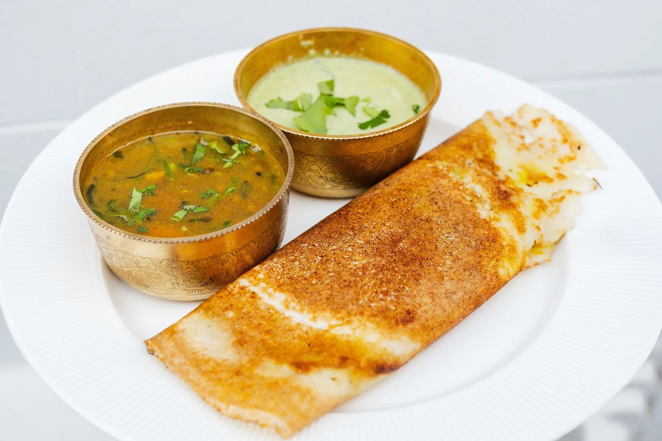 Crisp dosa on a white plate with two brass bowls of sambar and green chutney