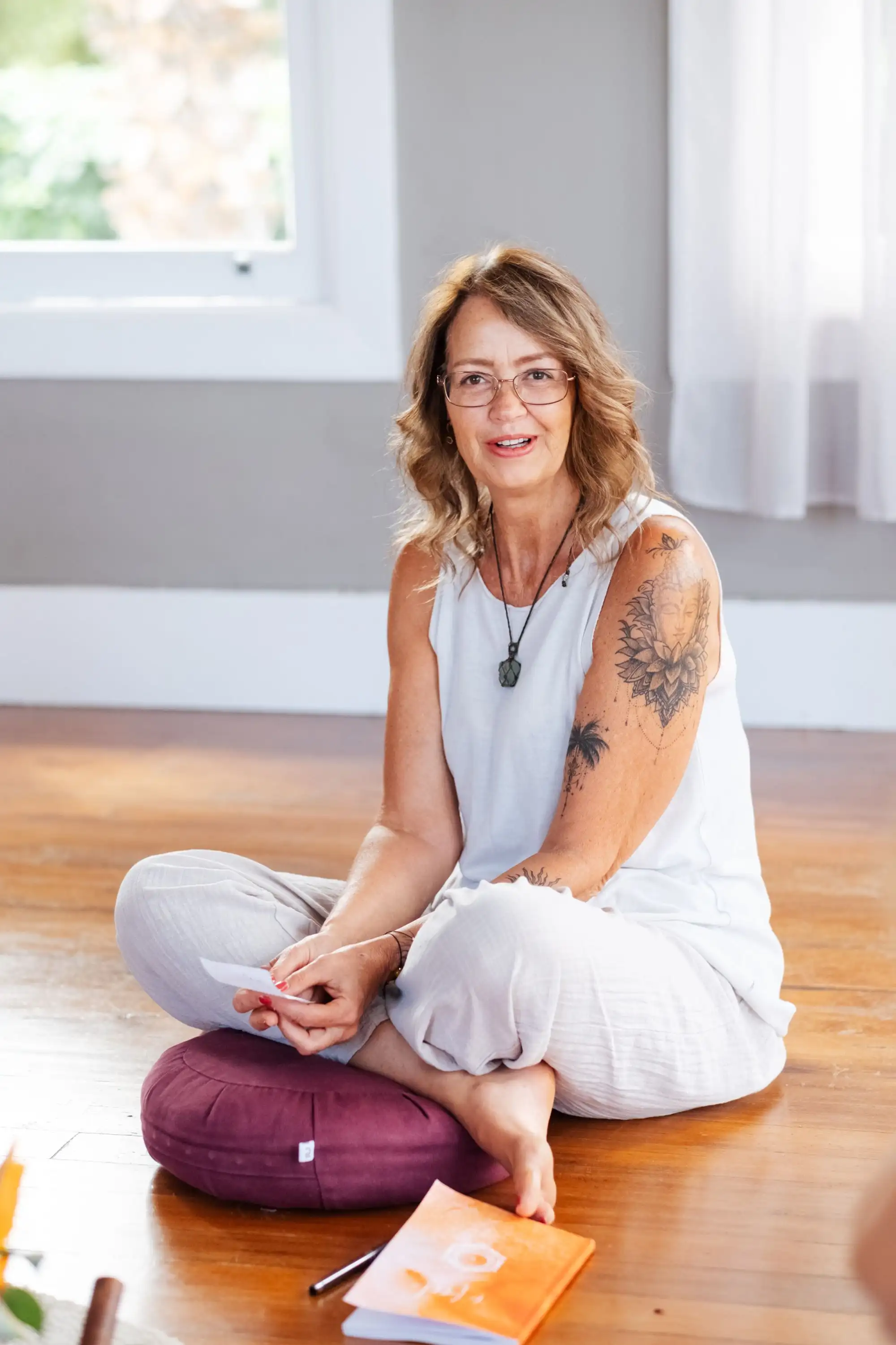 Smiling woman with glasses and tattooed arm sitting cross-legged on a cushion holding a card