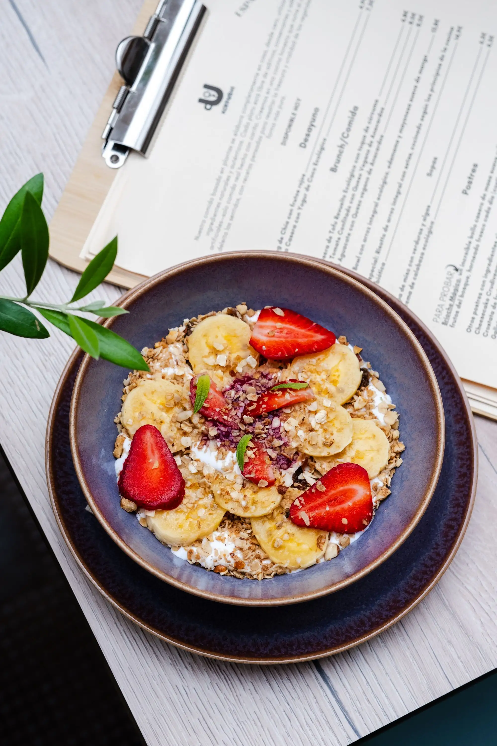 Bowl of granola with banana and strawberries beside a clipboard menu