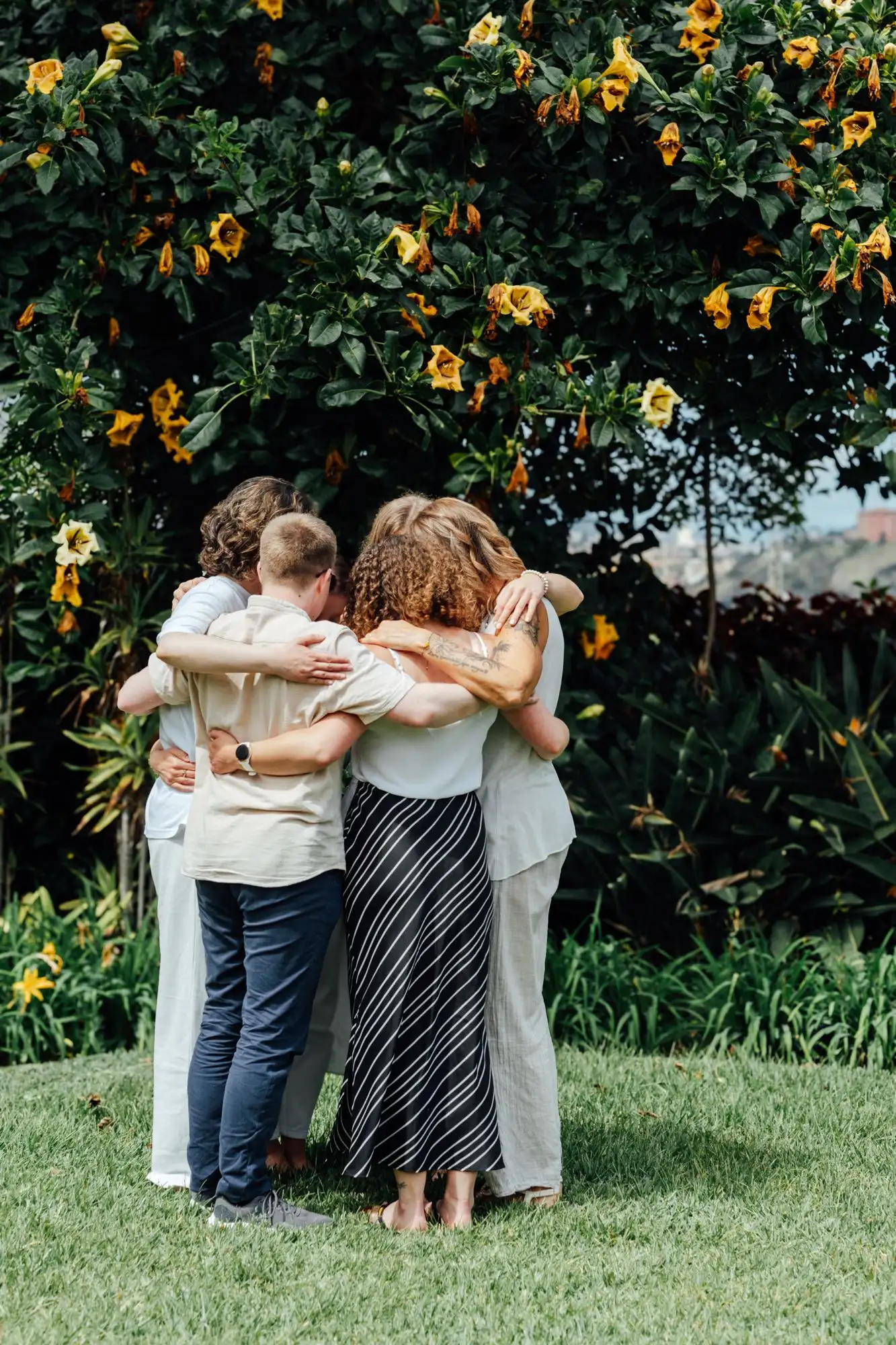 Group of people hugging in a circle on a lawn in front of a shrub with yellow flowers, seen from behind