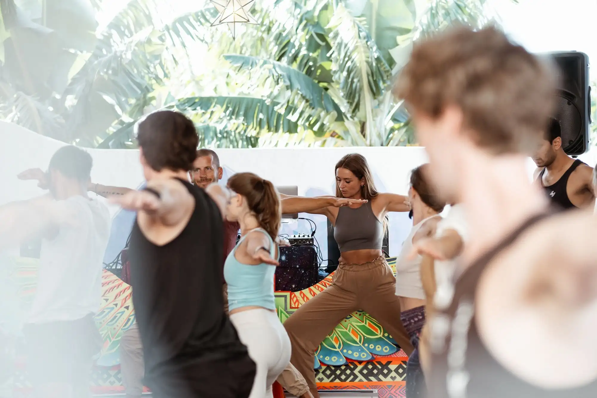 Group holding a wide-armed yoga pose on a sunny terrace with banana leaves and a colourful mat