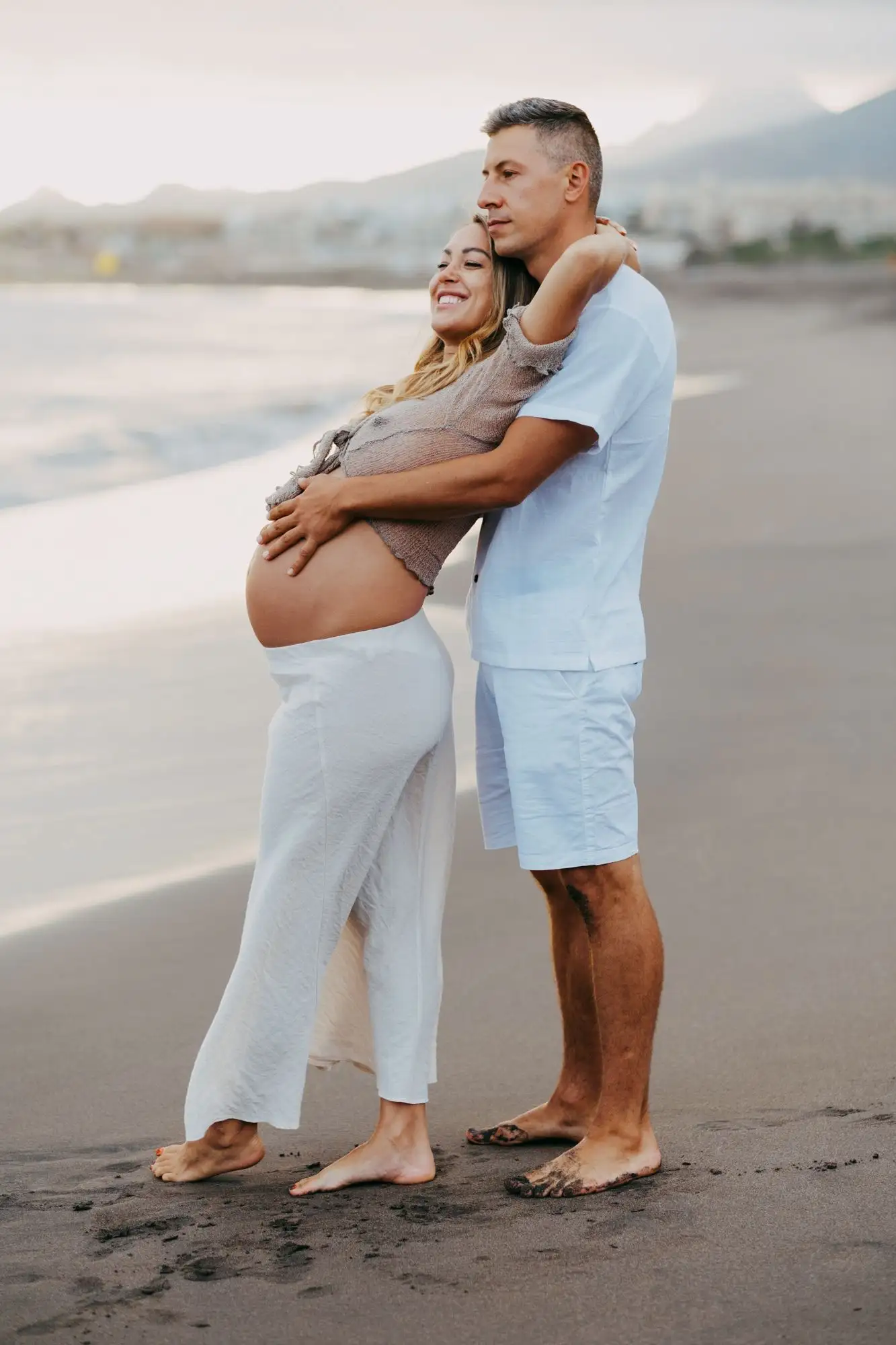 Smiling pregnant woman and man in white embracing on a black sand beach with mountains in the distance
