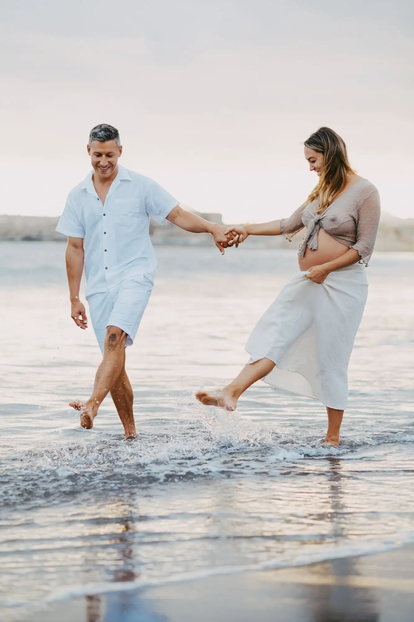 Smiling man and pregnant woman holding hands and splashing through shallow water on a beach