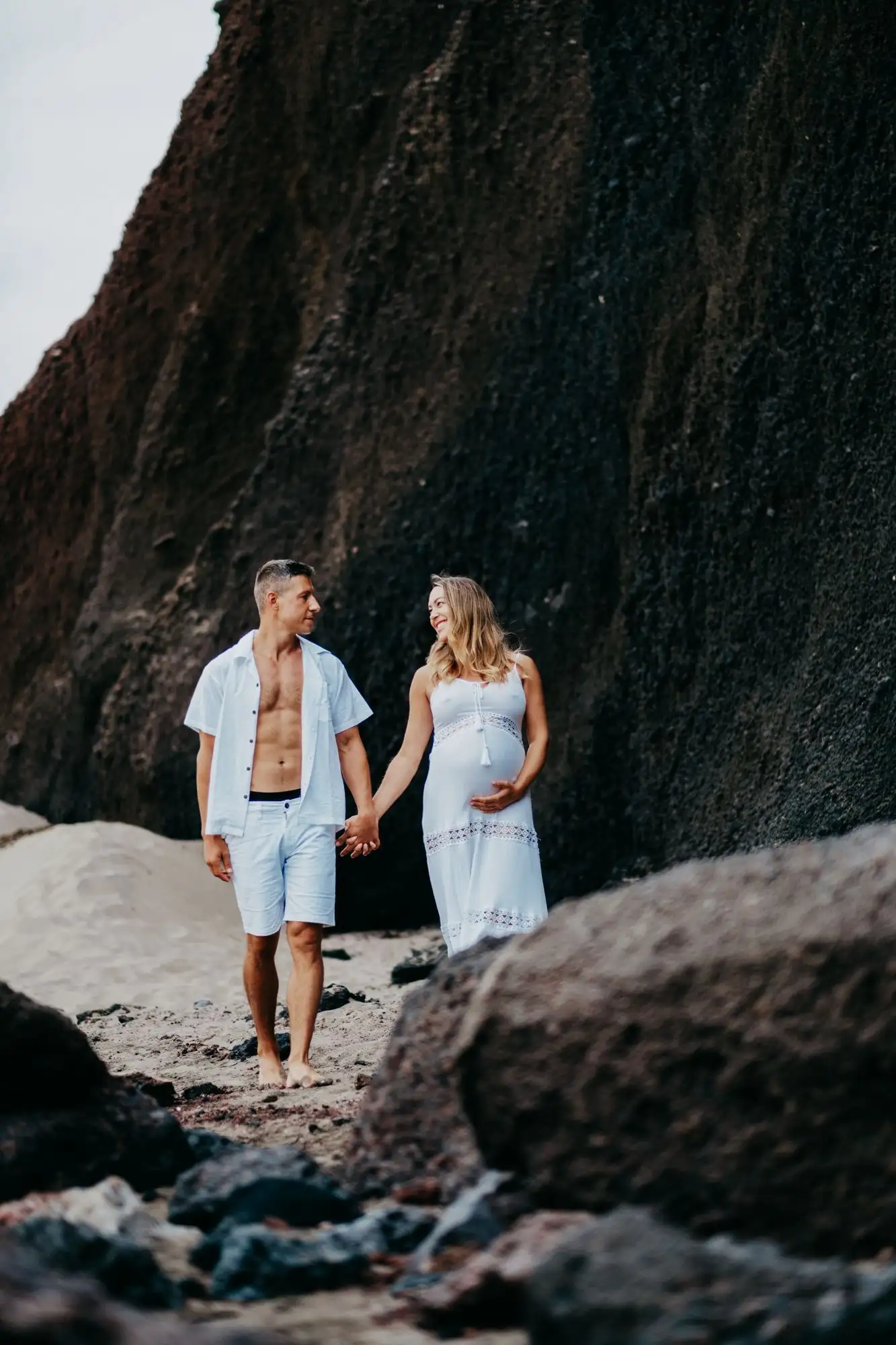 Pregnant woman in white and man in an open white shirt walking hand in hand below a dark volcanic rock face