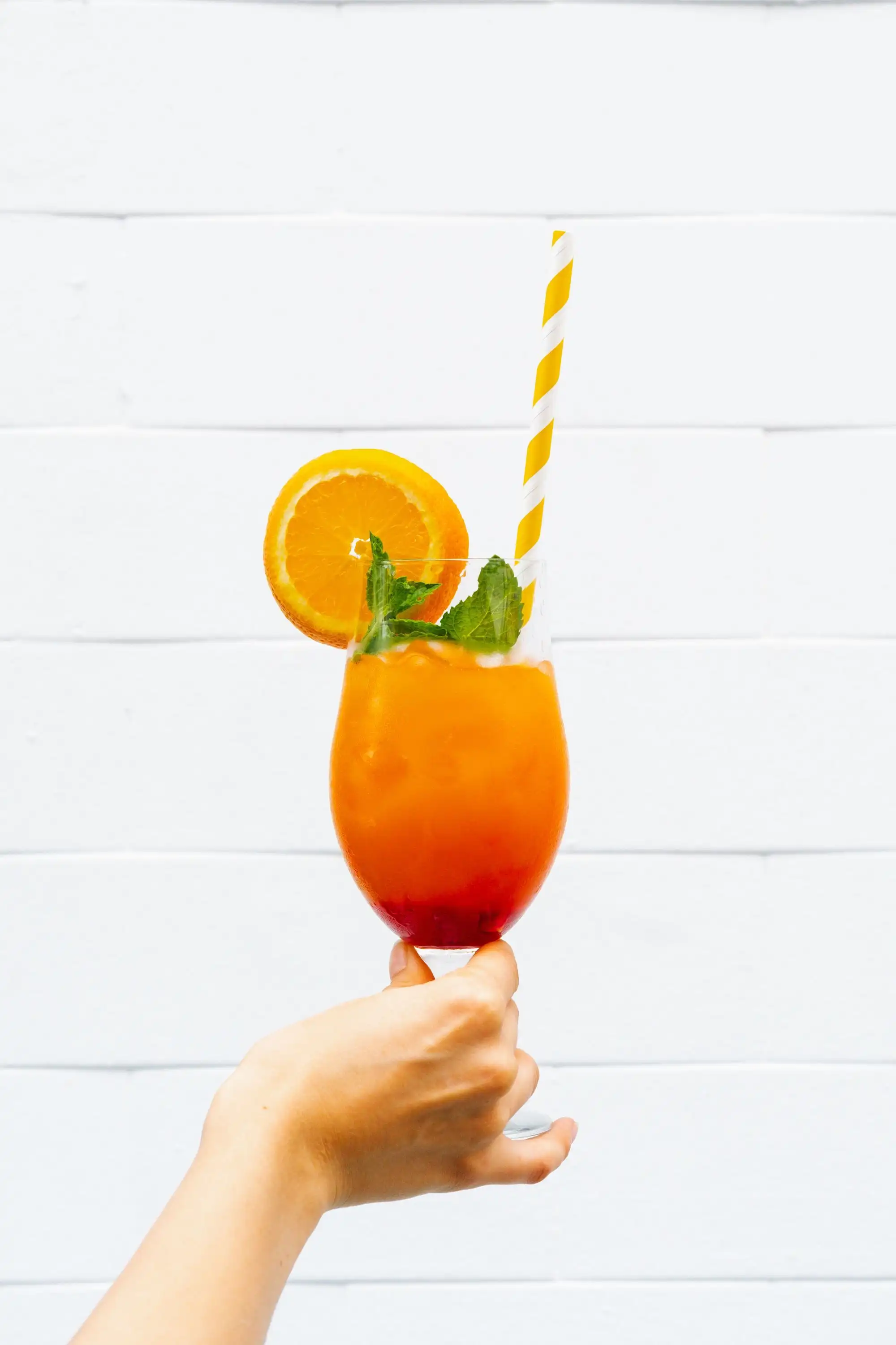 Hand holding an orange cocktail with a striped straw and orange slice against a white wall
