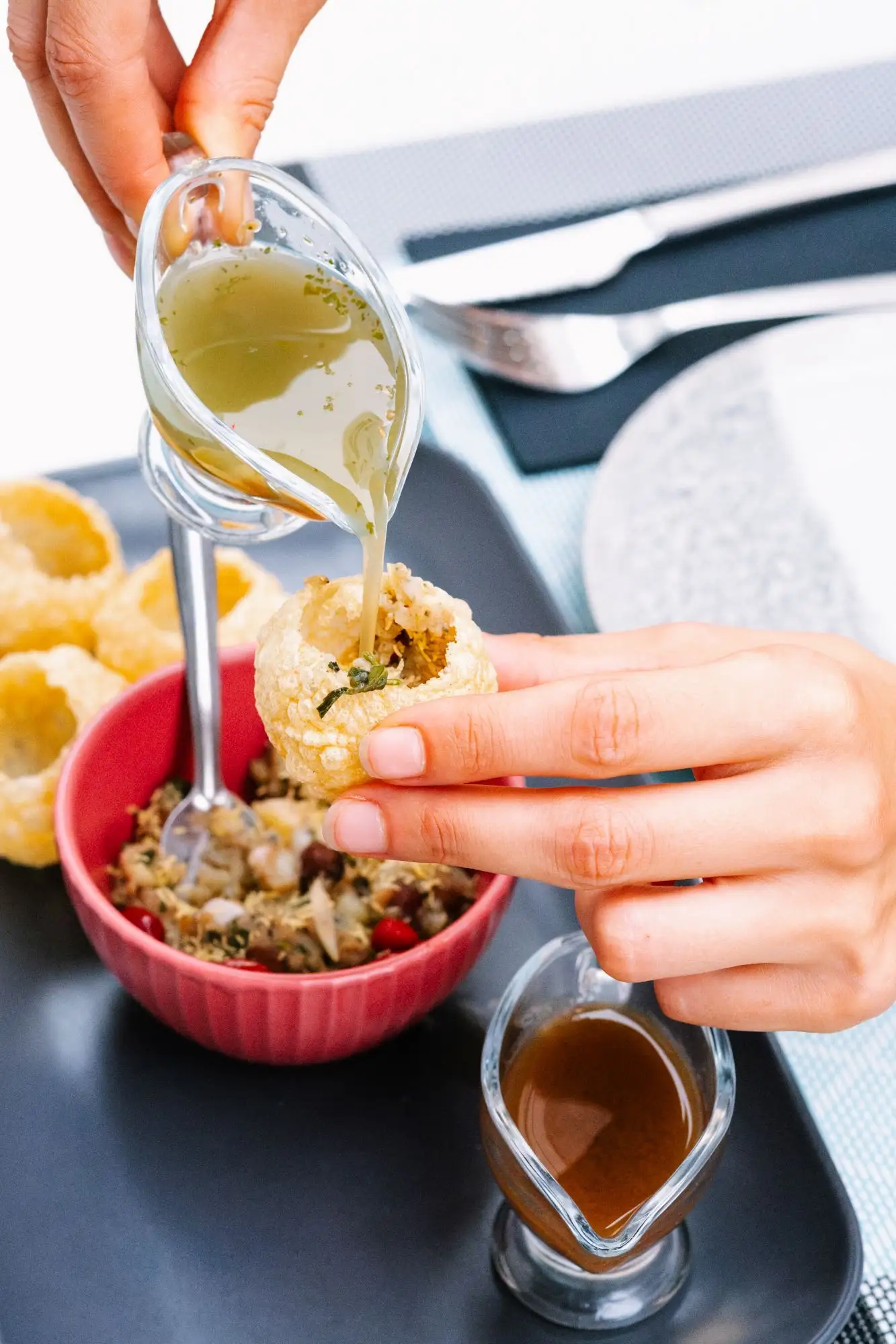 Hand pouring spiced water into a crisp puri above a bowl of filling