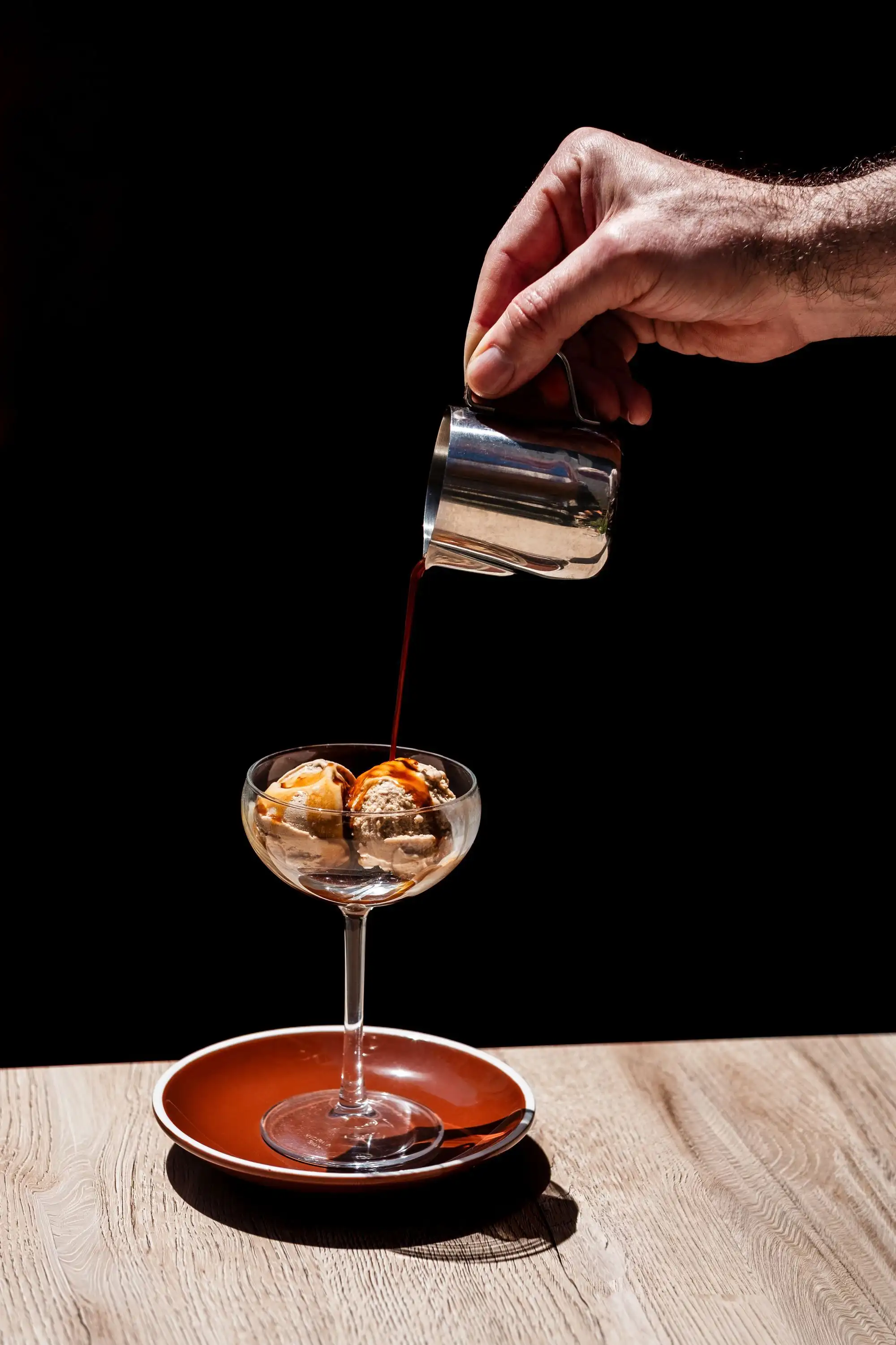 Hand pouring espresso from a steel jug over ice cream in a stemmed glass on a black background