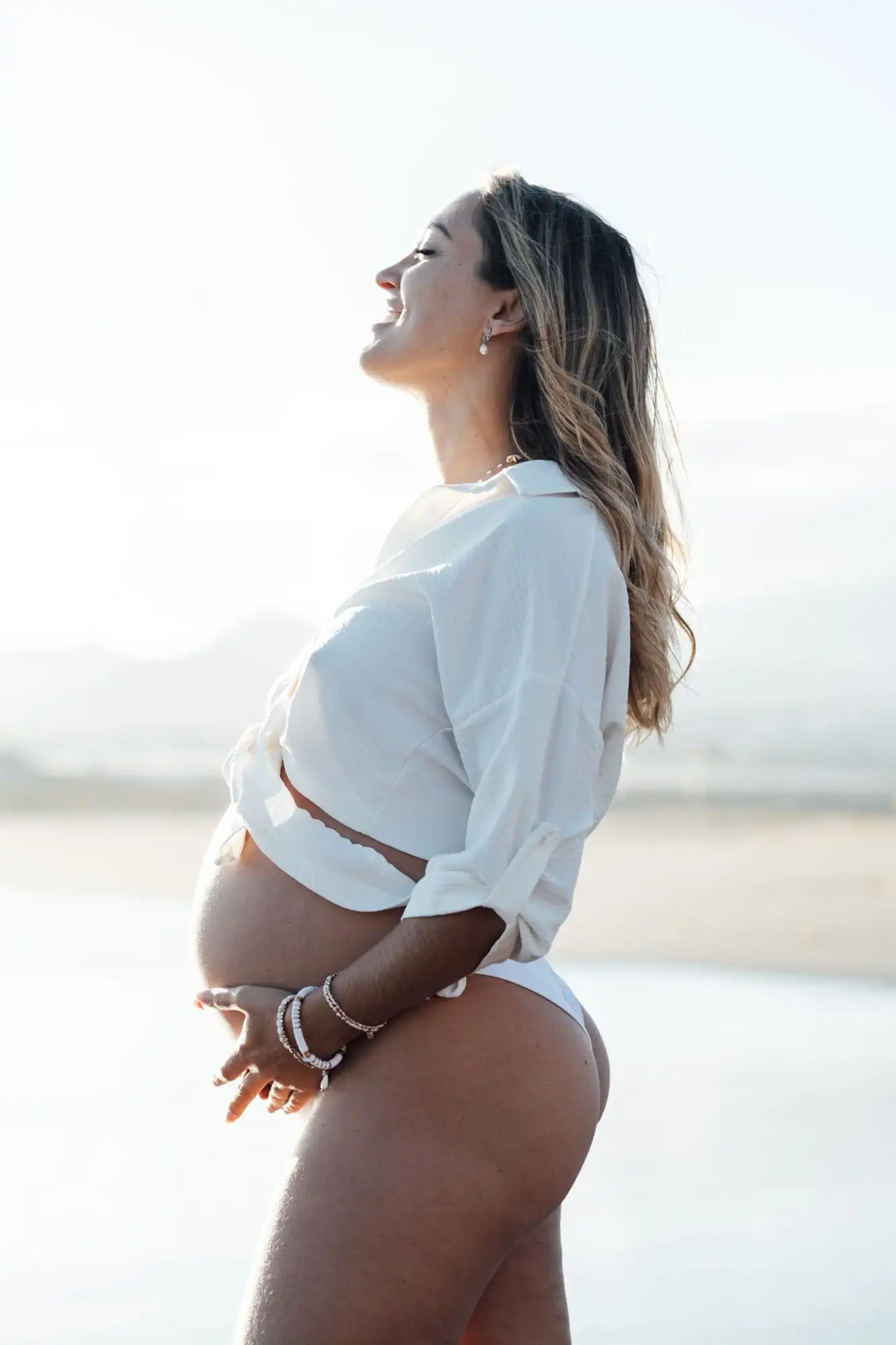 Pregnant woman in a white shirt laughing with her head tilted back, in soft backlight on a beach