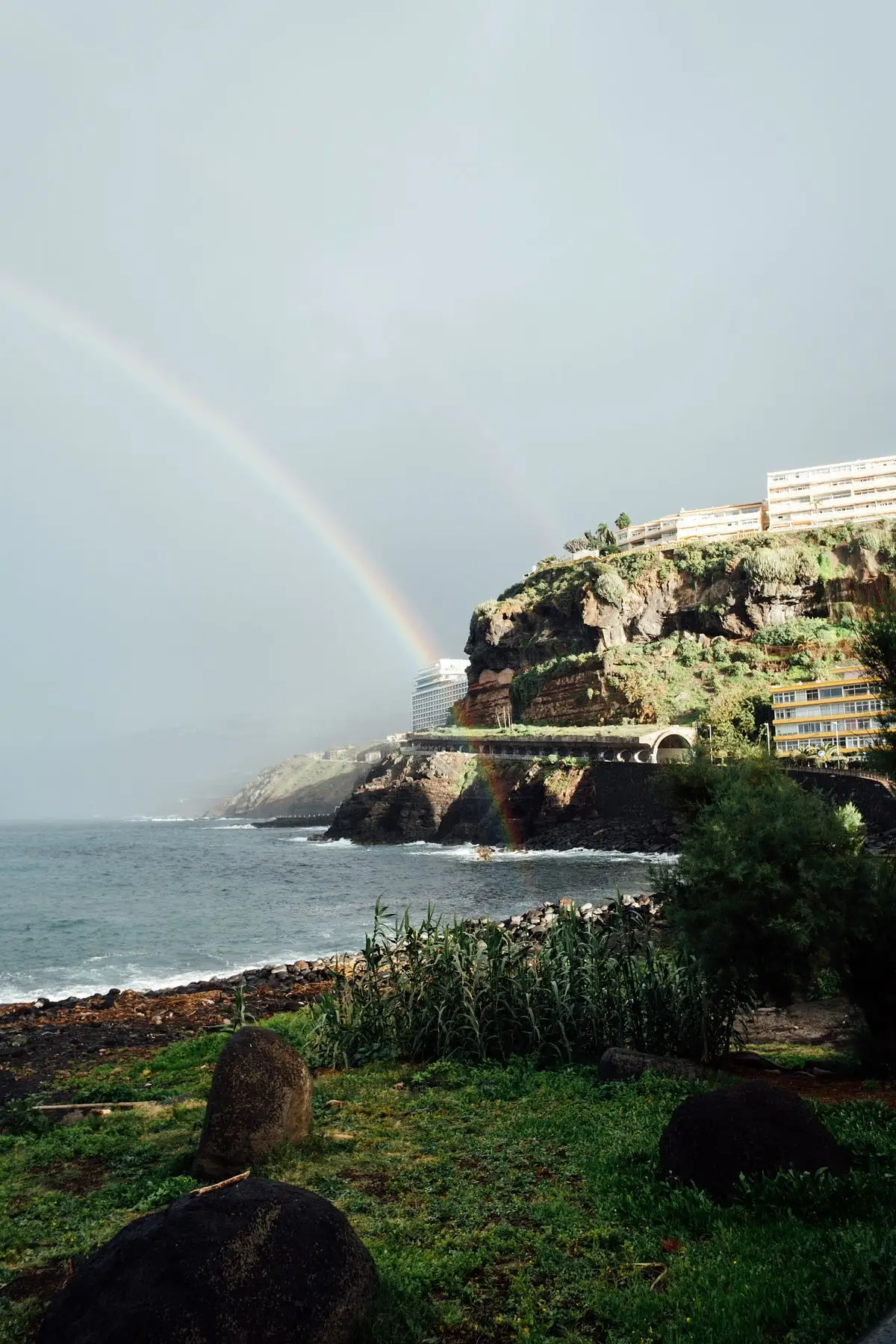 Arcoíris sobre un acantilado verde de la costa, con edificios de hotel y un mar gris