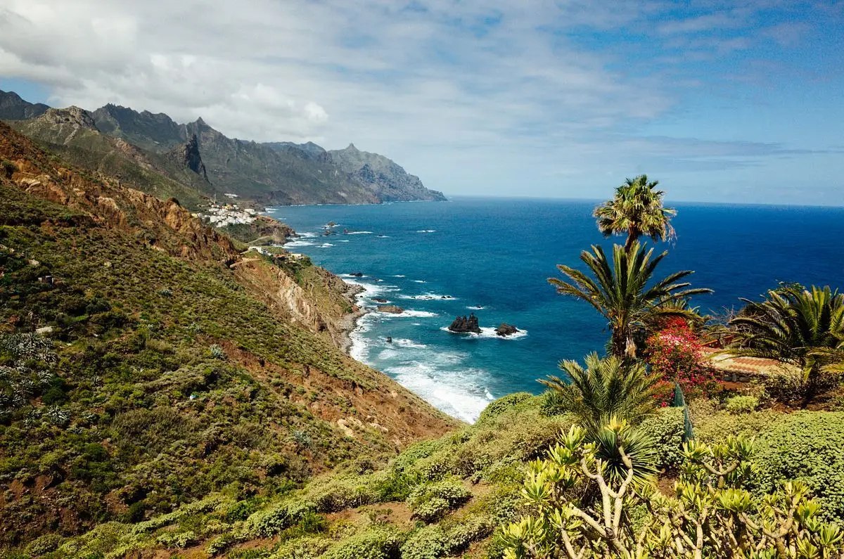 Costa escarpada con un pueblo blanco, roques, palmeras y un océano azul intenso bajo montañas afiladas