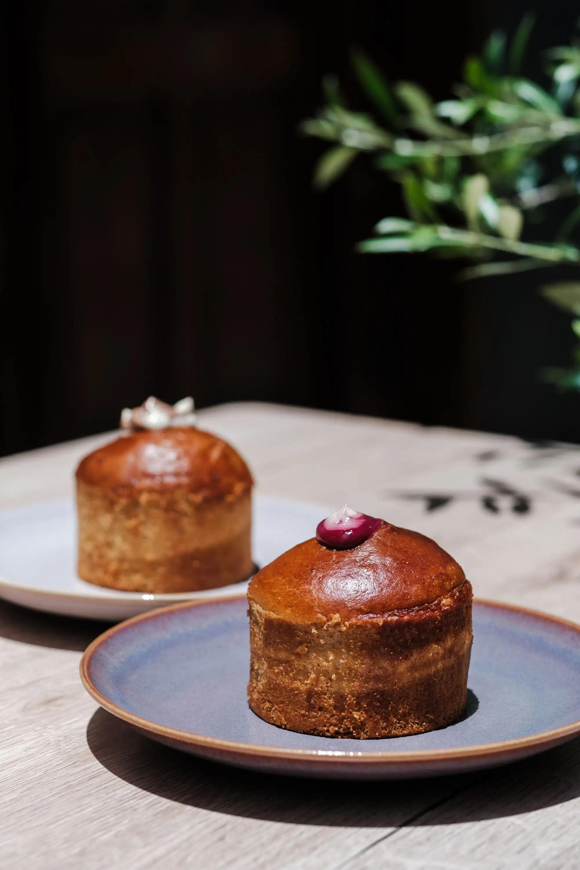 Dos pasteles glaseados pequeños en platos sobre una mesa, con una rama de olivo al fondo