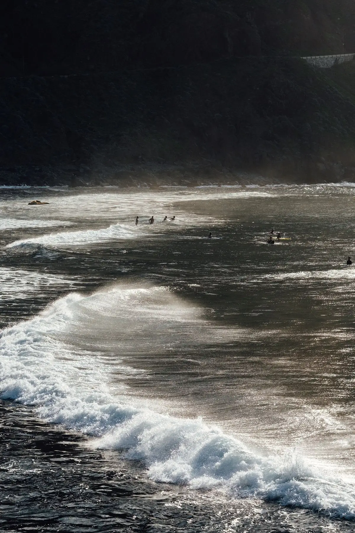 Olas rompiendo en una playa oscura con bañistas diminutos bajo un acantilado escarpado