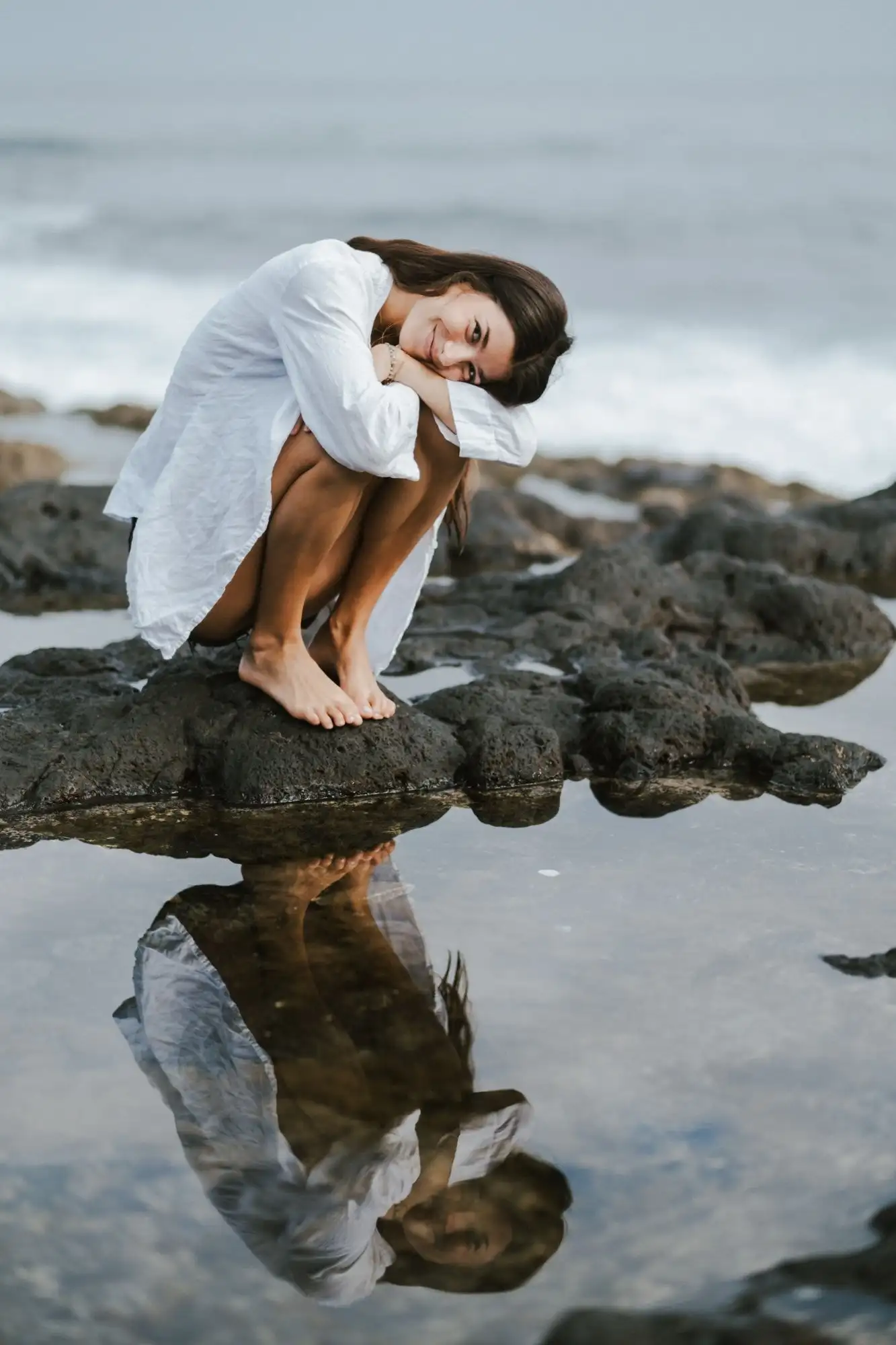 Woman in a white shirt crouching on a black rock with her head on her knees, reflected in a still tide pool
