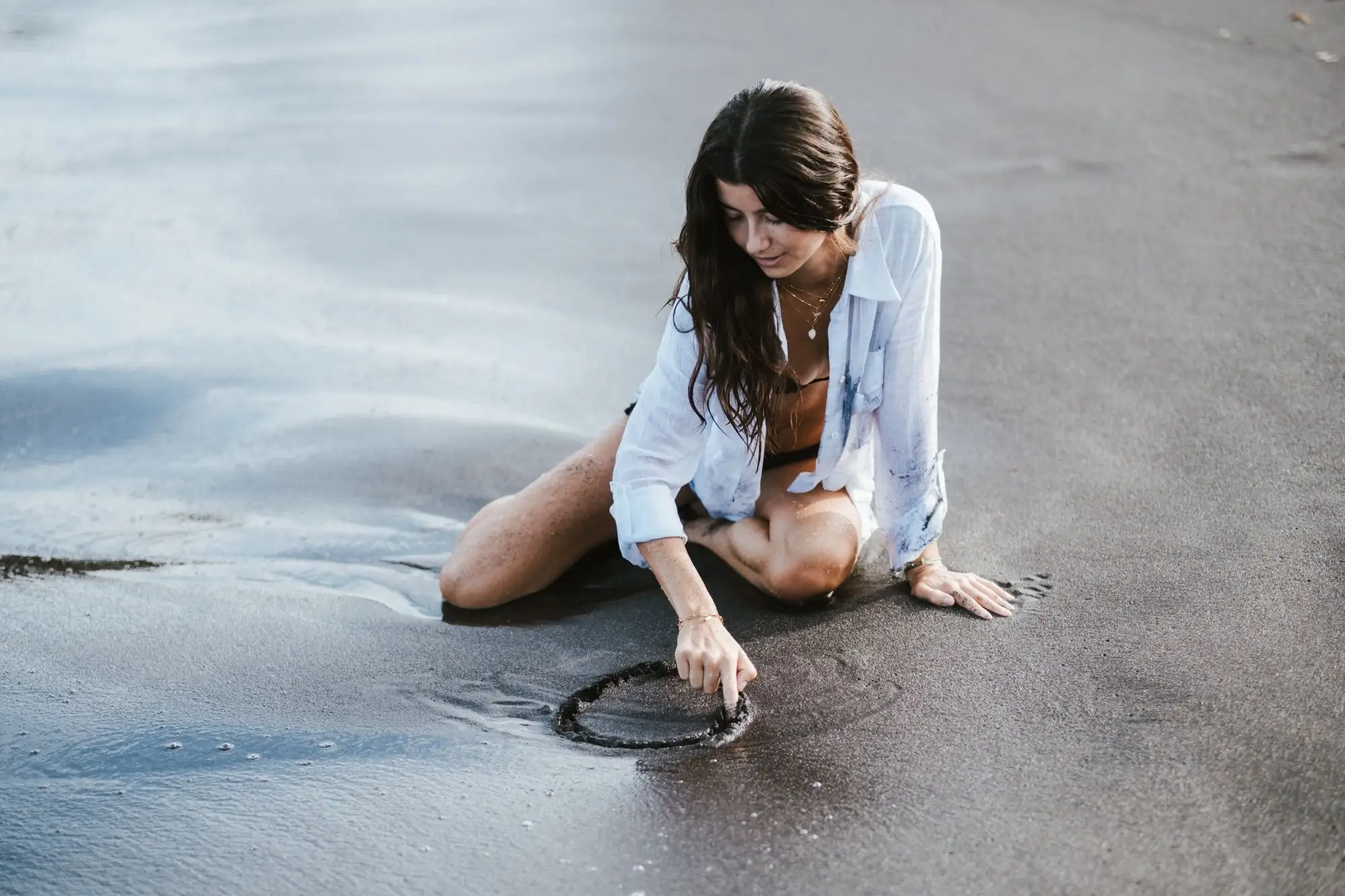 Woman in a white shirt kneeling on dark wet sand, drawing a circle in the sand with her finger