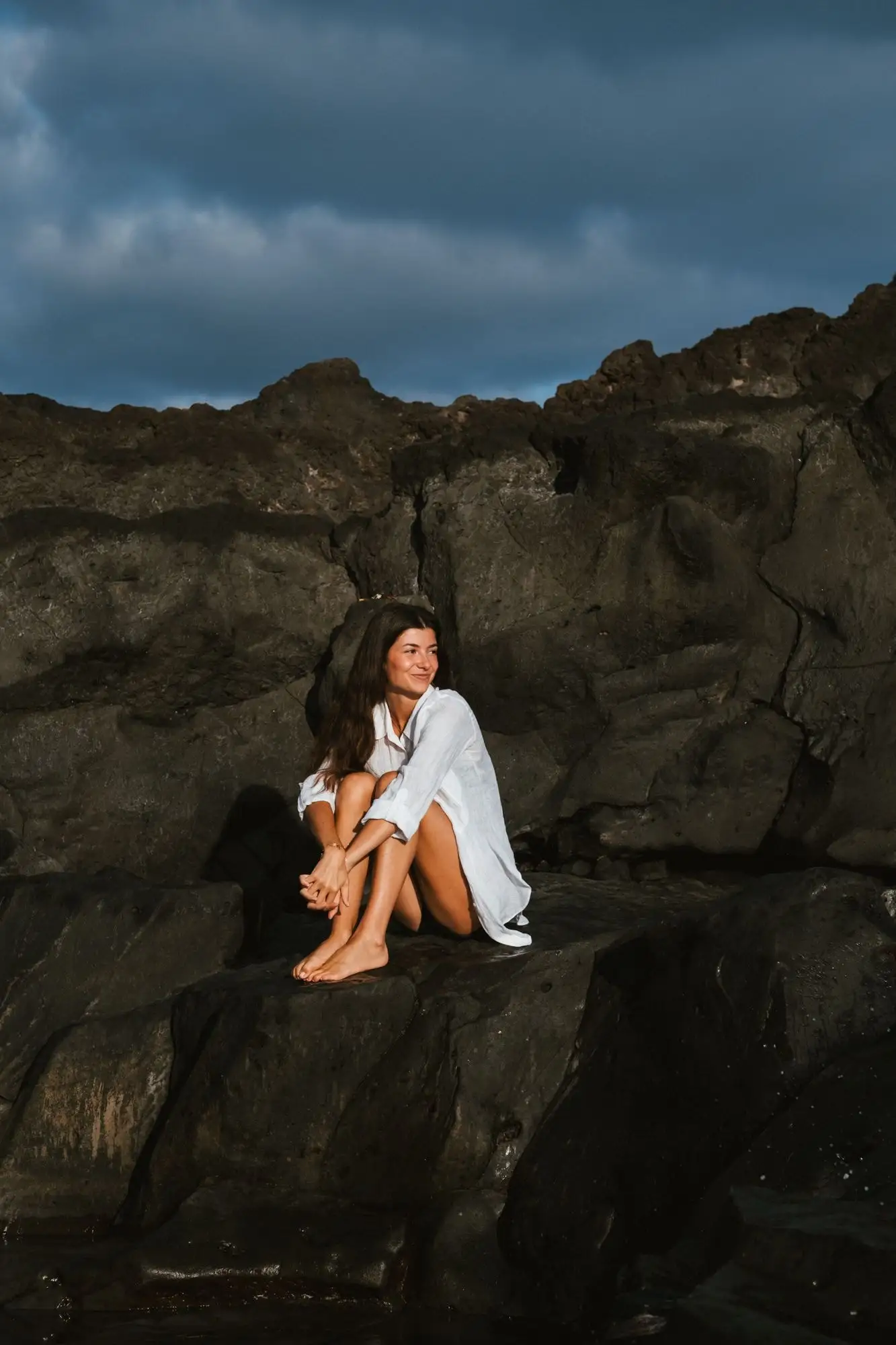 Mujer con camisa blanca abrazándose las rodillas sobre una roca de lava oscura bajo un cielo azul tormentoso