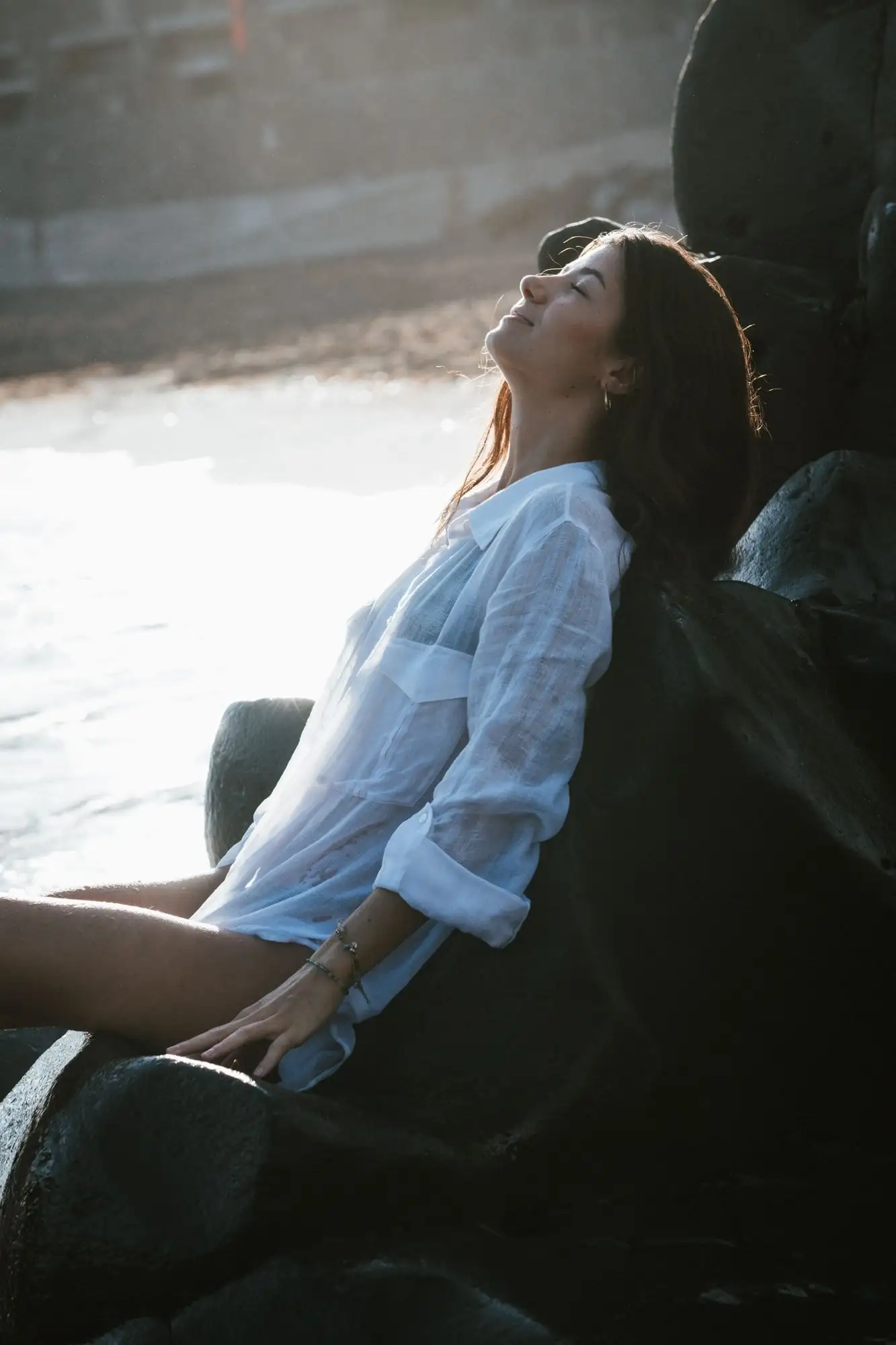 Woman leaning back against black rocks with her face lifted to the sun, white shirt, bright sea glare behind
