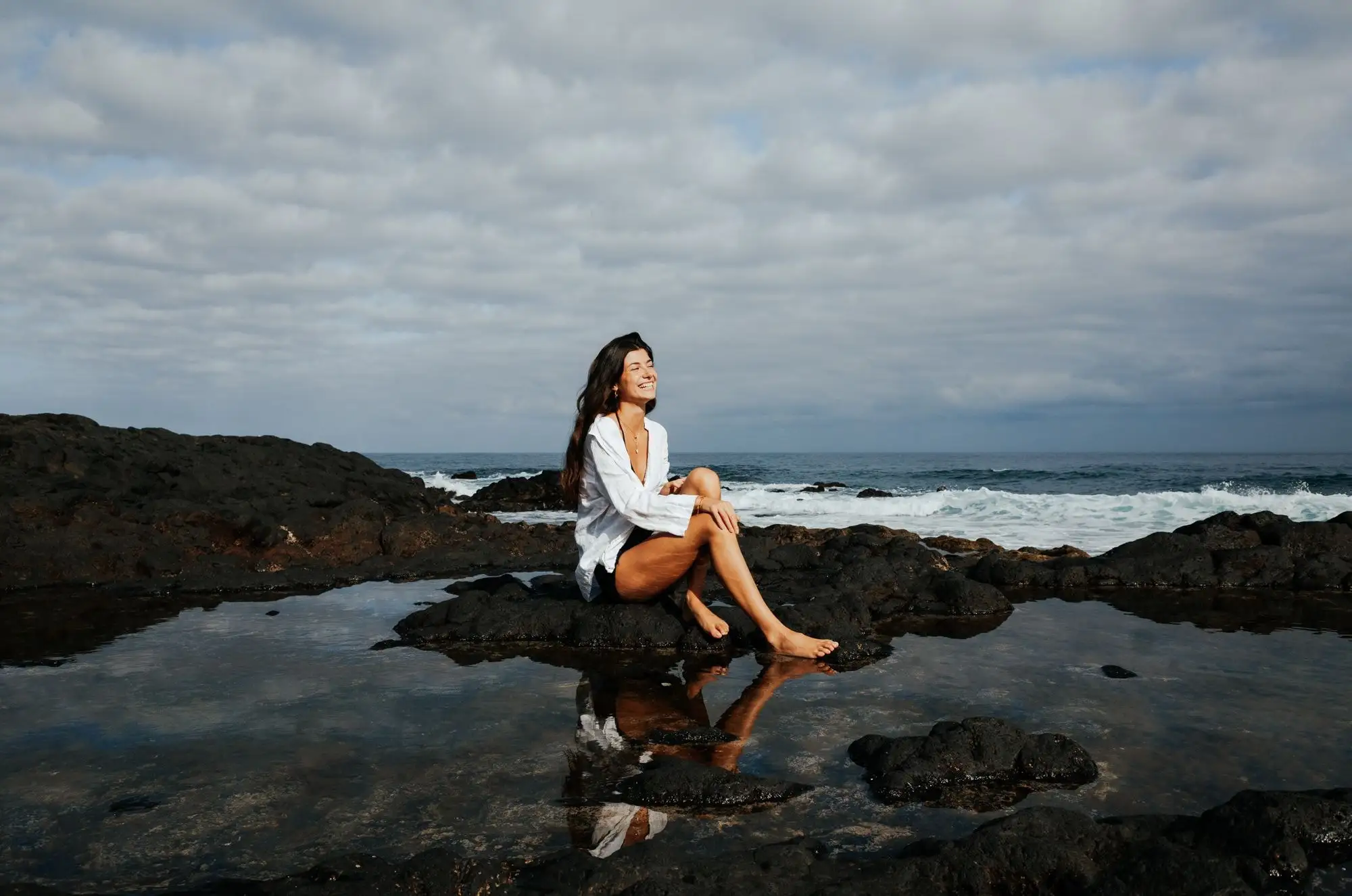 Smiling woman sitting on a rock beside a still tide pool that mirrors her, under heavy clouds