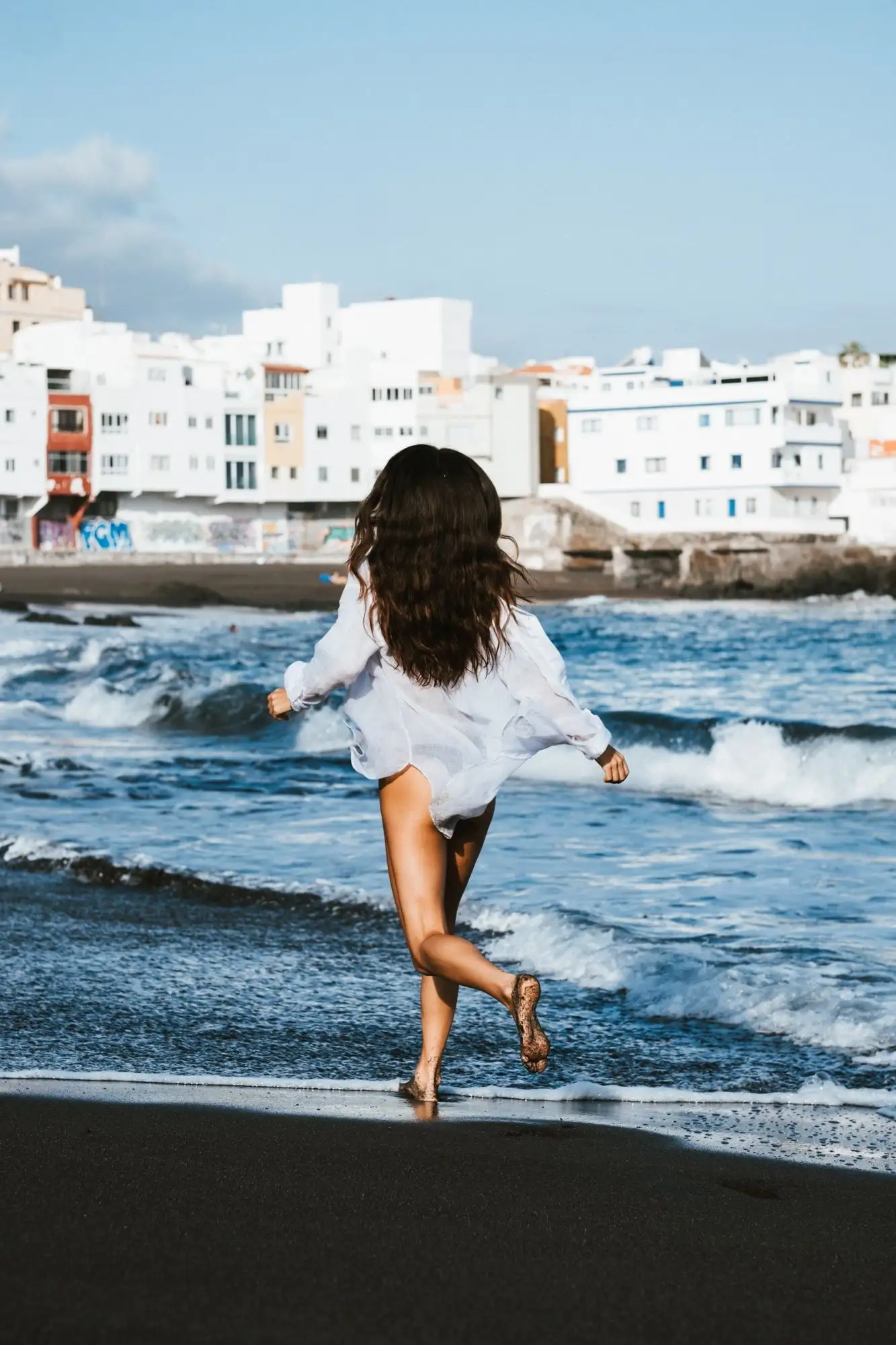 Woman in a white shirt running into the sea on a black-sand beach, seen from behind, white village in the distance