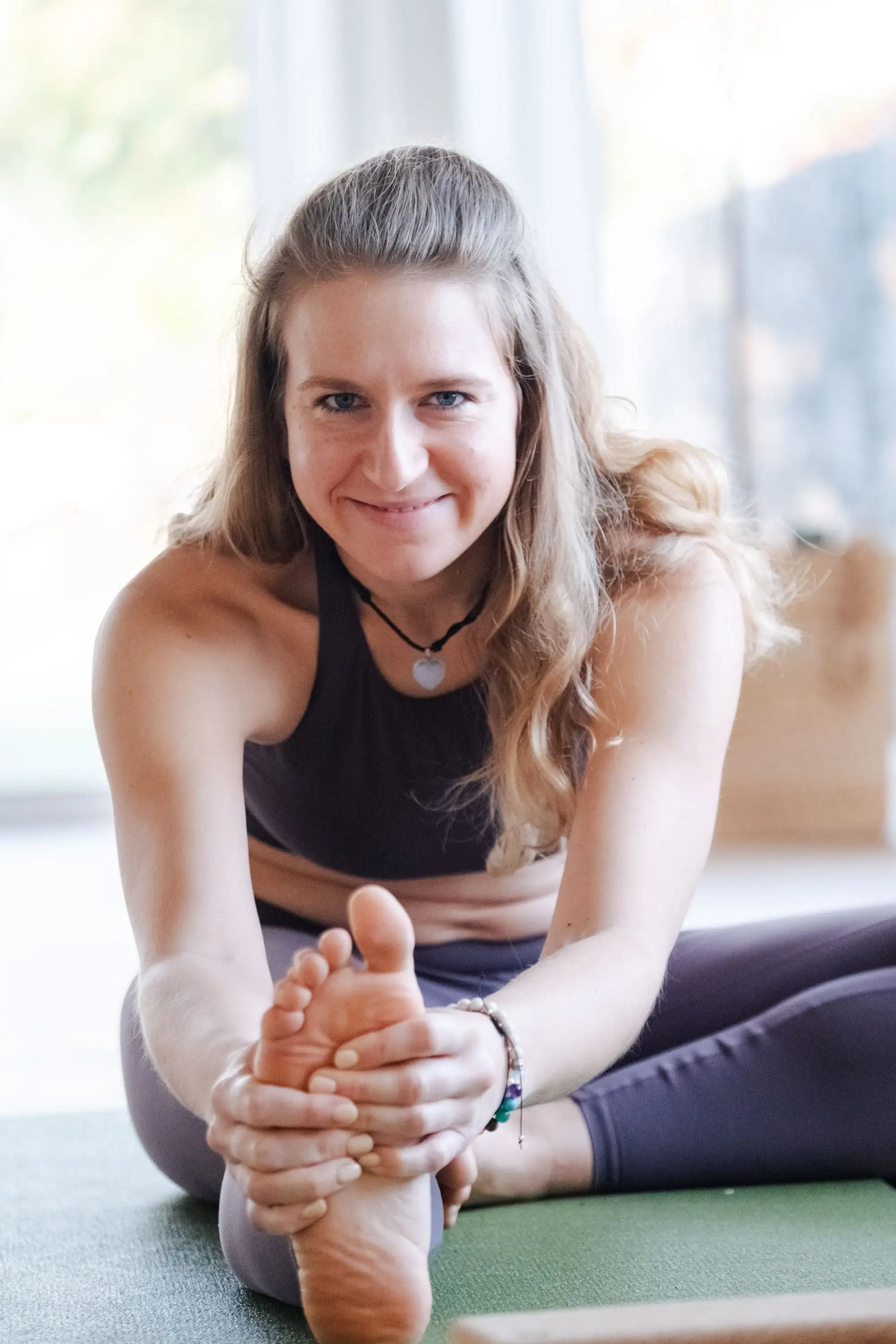 Smiling woman seated on a yoga mat holding her foot in a seated forward fold