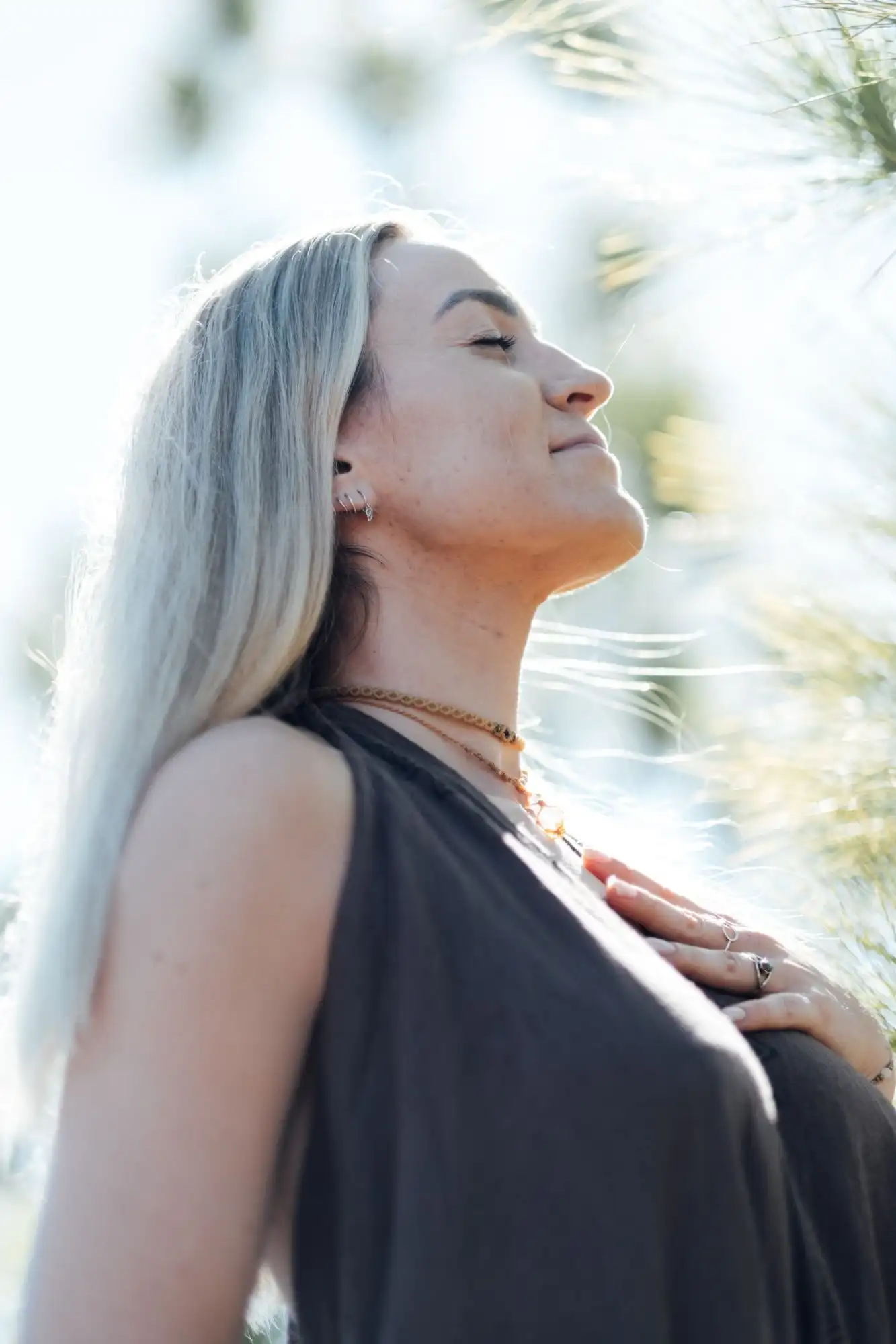 Woman with silver-blonde hair and eyes closed, hand on her chest, backlit among pine needles