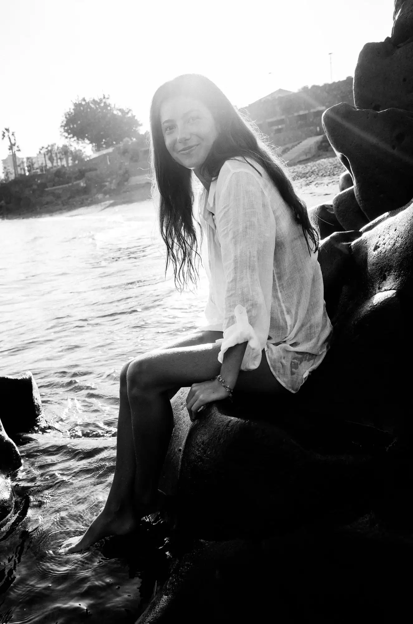 Black and white portrait of a woman with long hair sitting on lava rocks by the sea in bright backlight