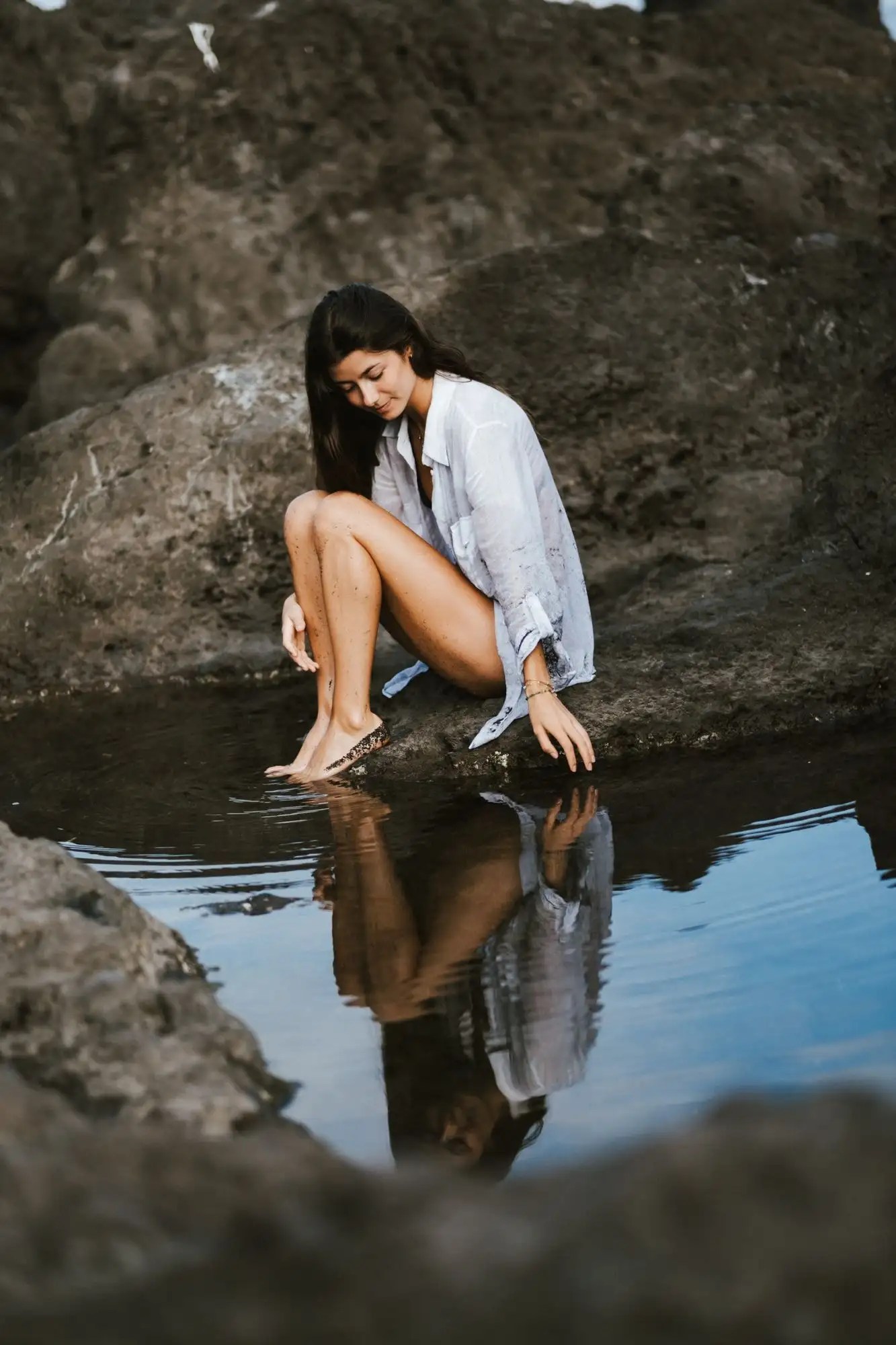 Woman in a white shirt sitting on a rock and dipping her foot toward a tide pool that reflects her