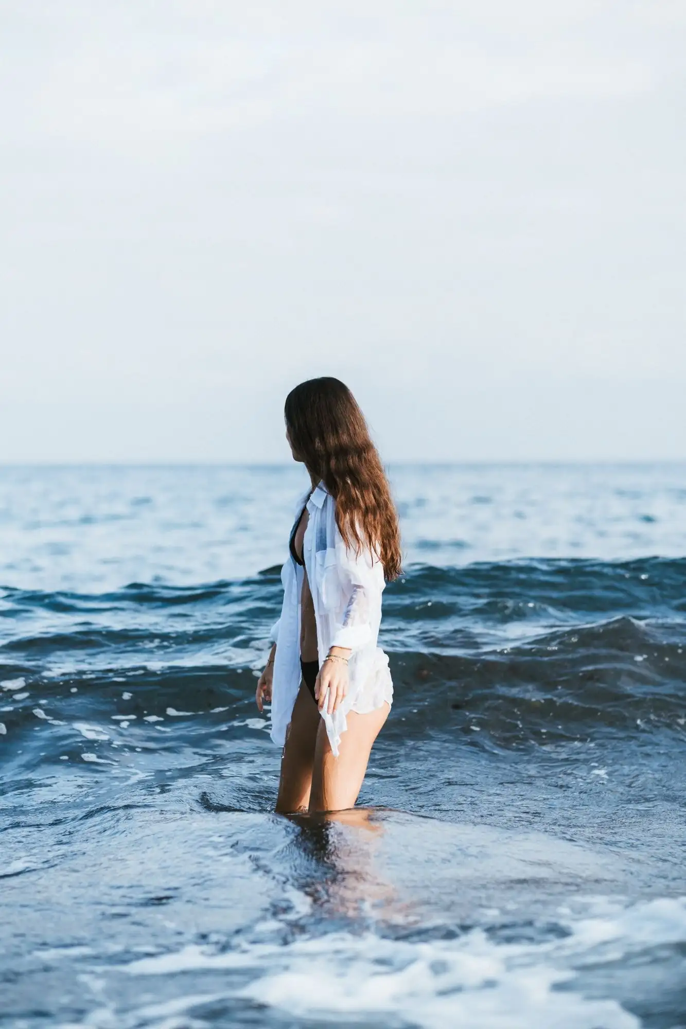 Woman with long hair standing knee-deep in blue waves, looking away in a white shirt