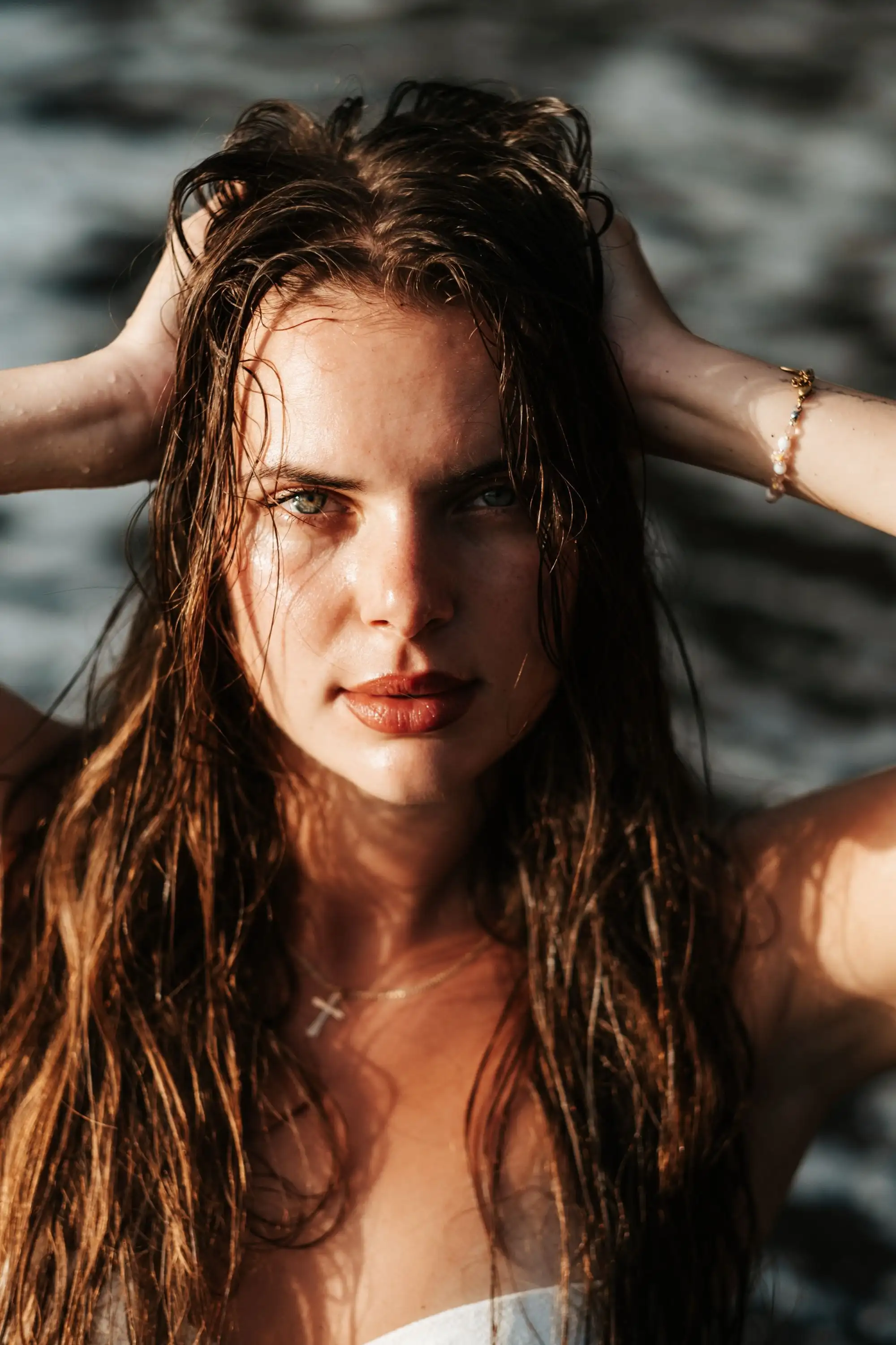 Close portrait of a woman with wet hair, hands in her hair, looking straight into the camera, sea behind