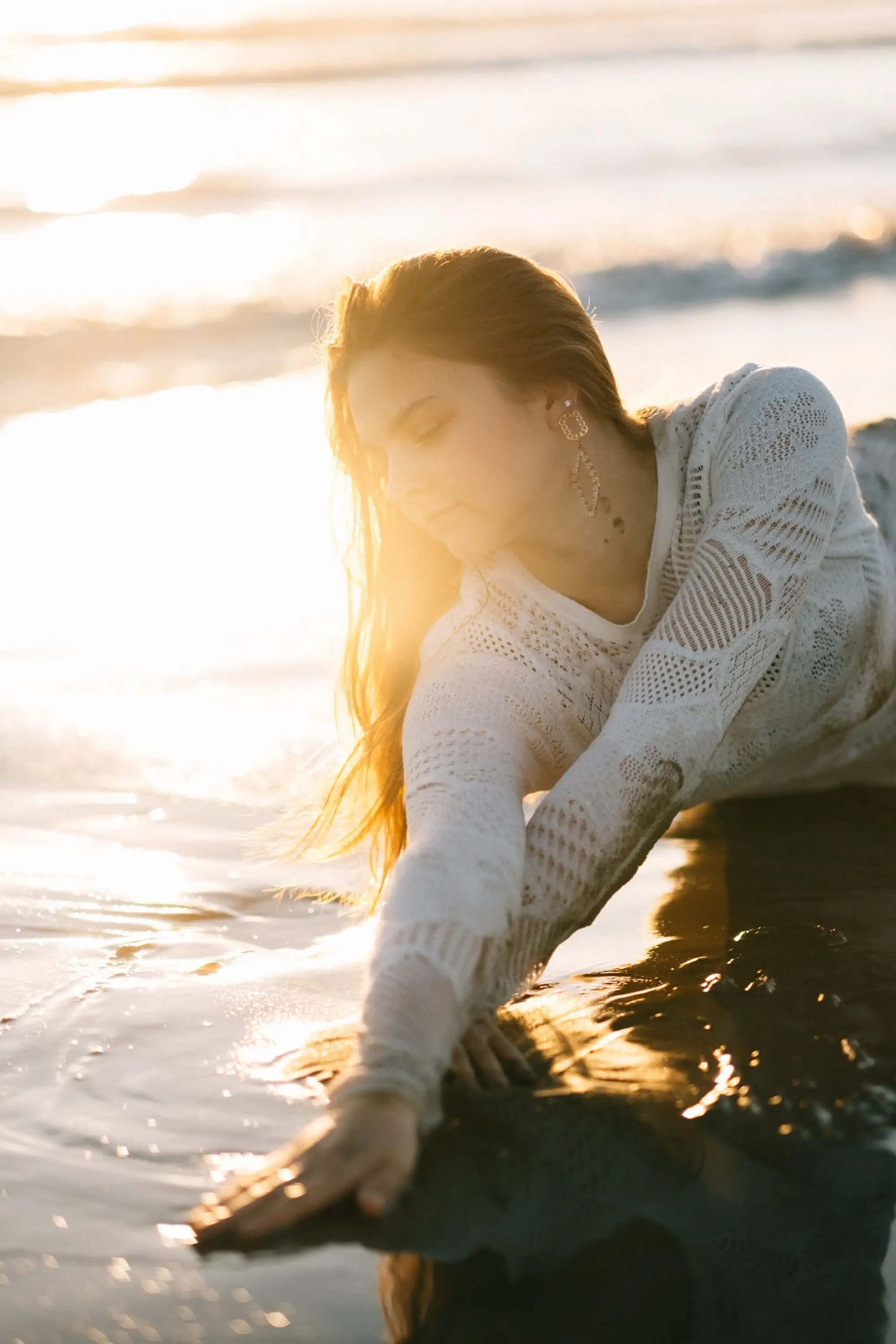 Woman in a white crochet top leaning on dark rock with her hand in sunlit, rippling water