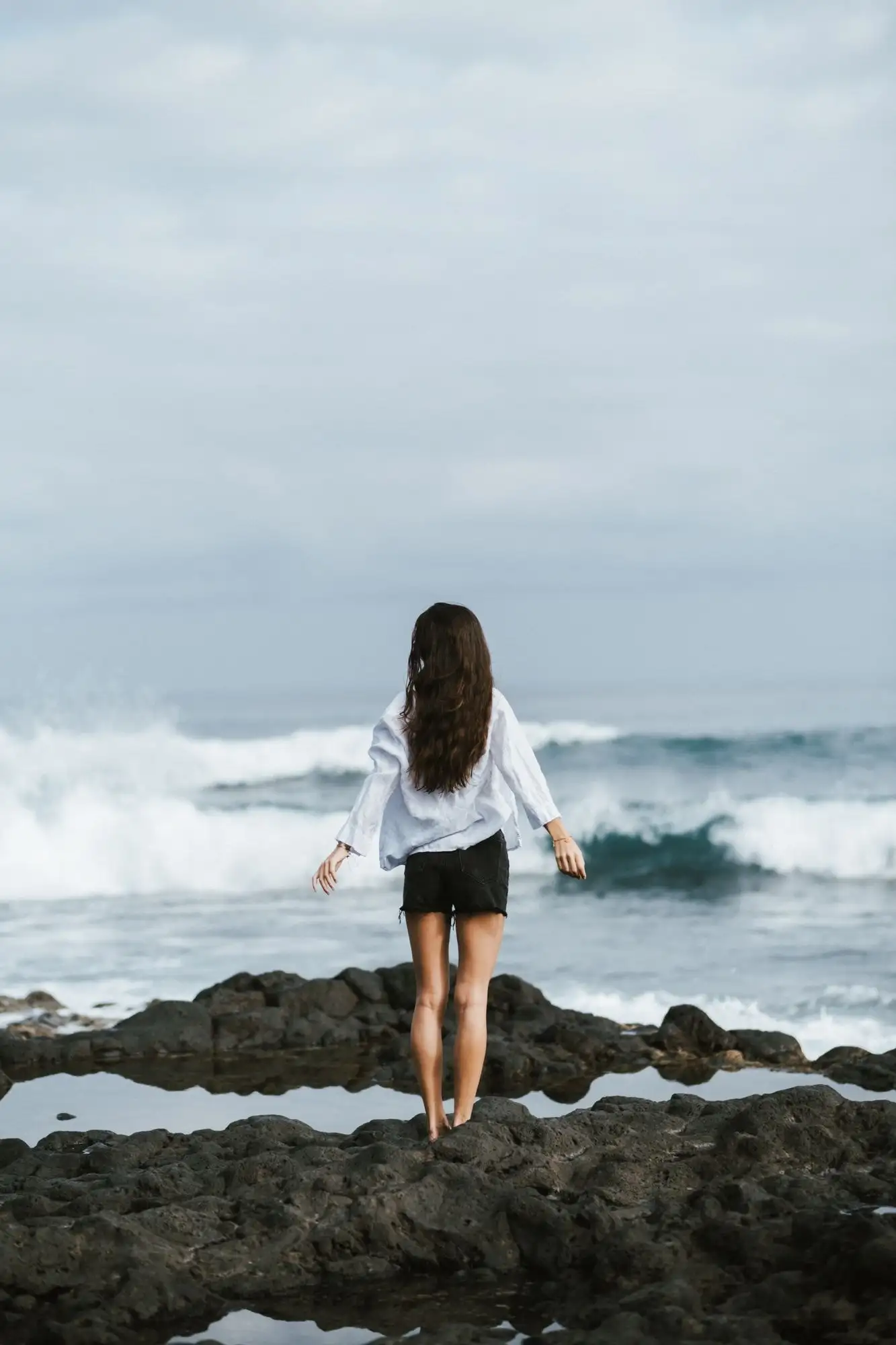 Woman in white shirt and black shorts seen from behind, standing on rocks facing breaking waves